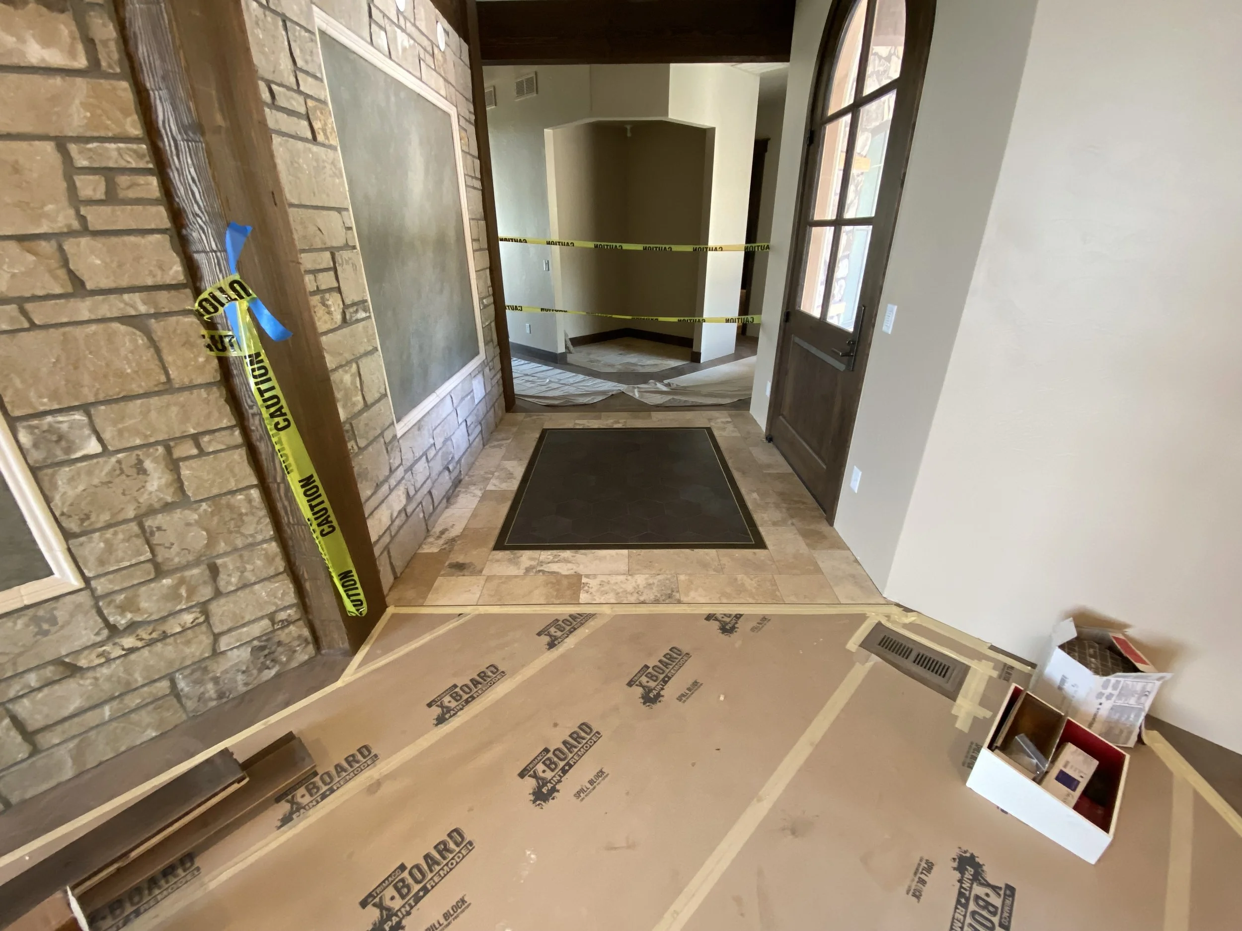 Interior view of a house under construction with caution tape across the doorway, a black welcome mat on the tiled floor, partially finished walls, and building supplies nearby.