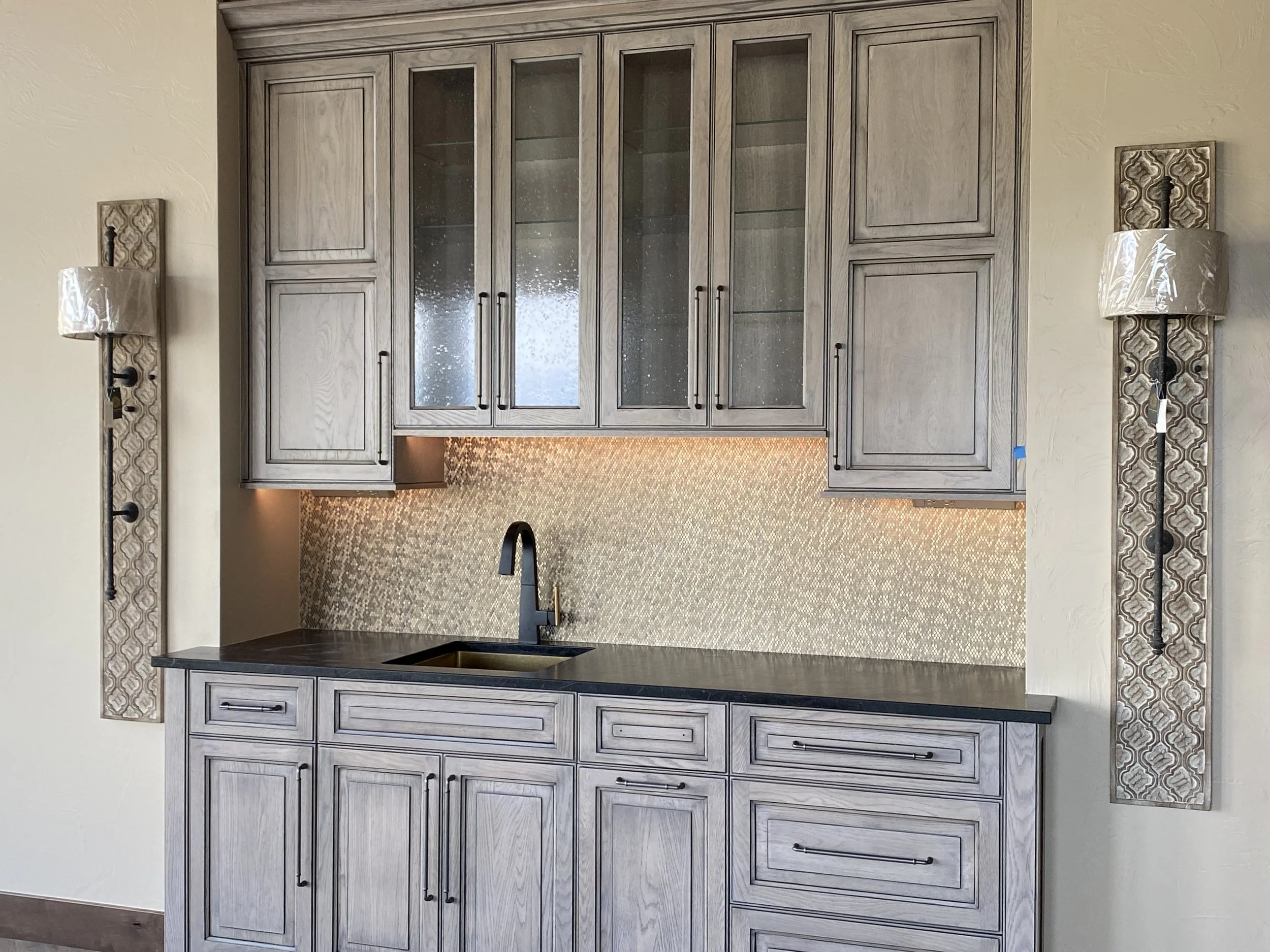 Kitchen area with gray wooden cabinets, a black countertop, a black sink with a black faucet, and a textured backsplash with under-cabinet lighting; wall-mounted lamps on either side of the kitchen.