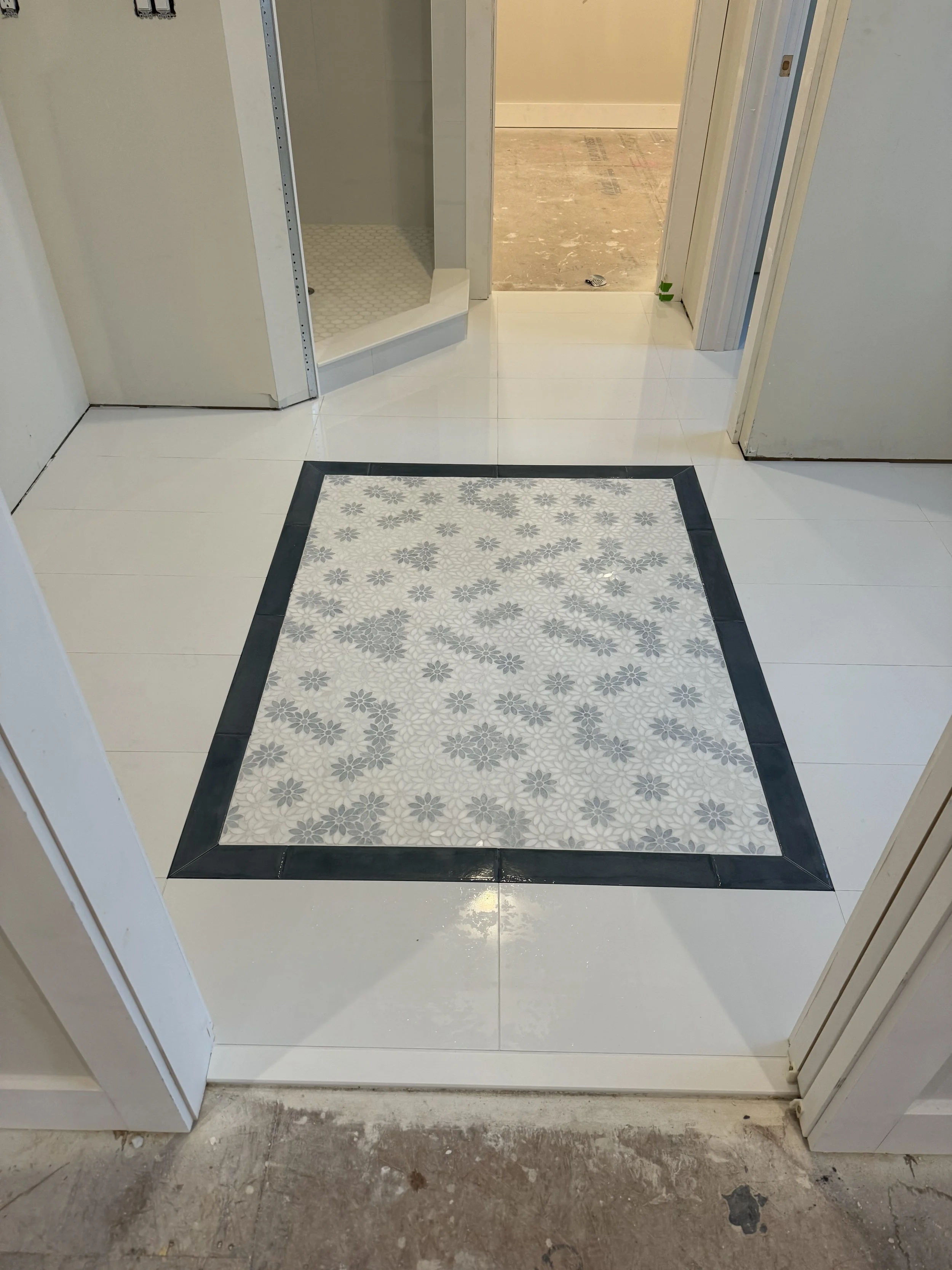 Clean bathroom or shower area with white tiled floor, decorative tile border, and a shower stall with a glass door in the background.