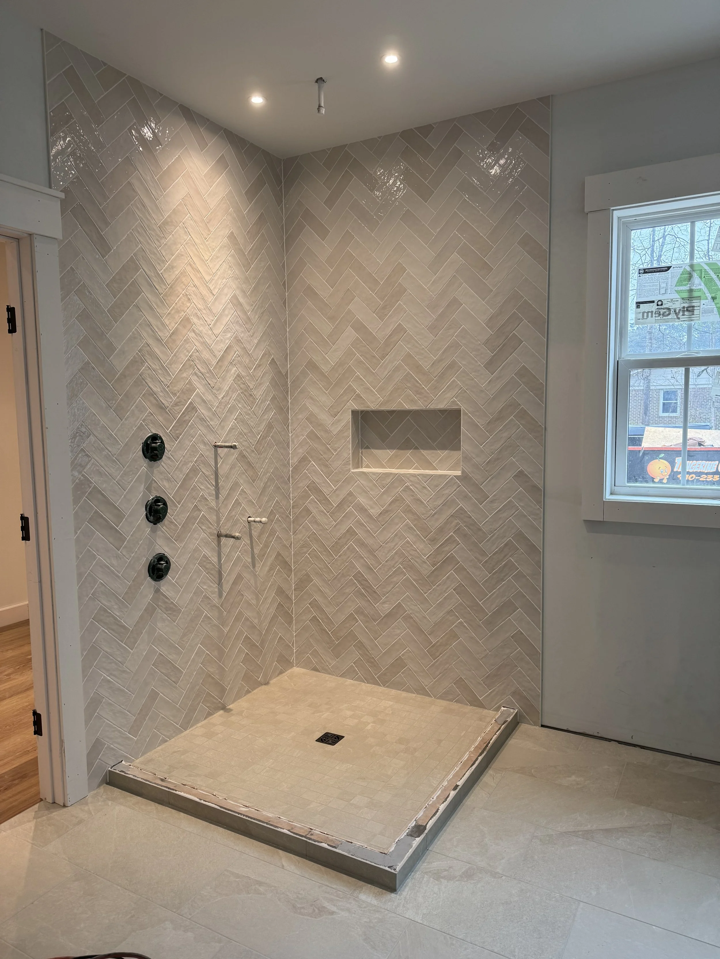 Unfinished shower area in a bathroom with beige herringbone tile walls, a built-in niche, and a tiled shower base, next to a window.