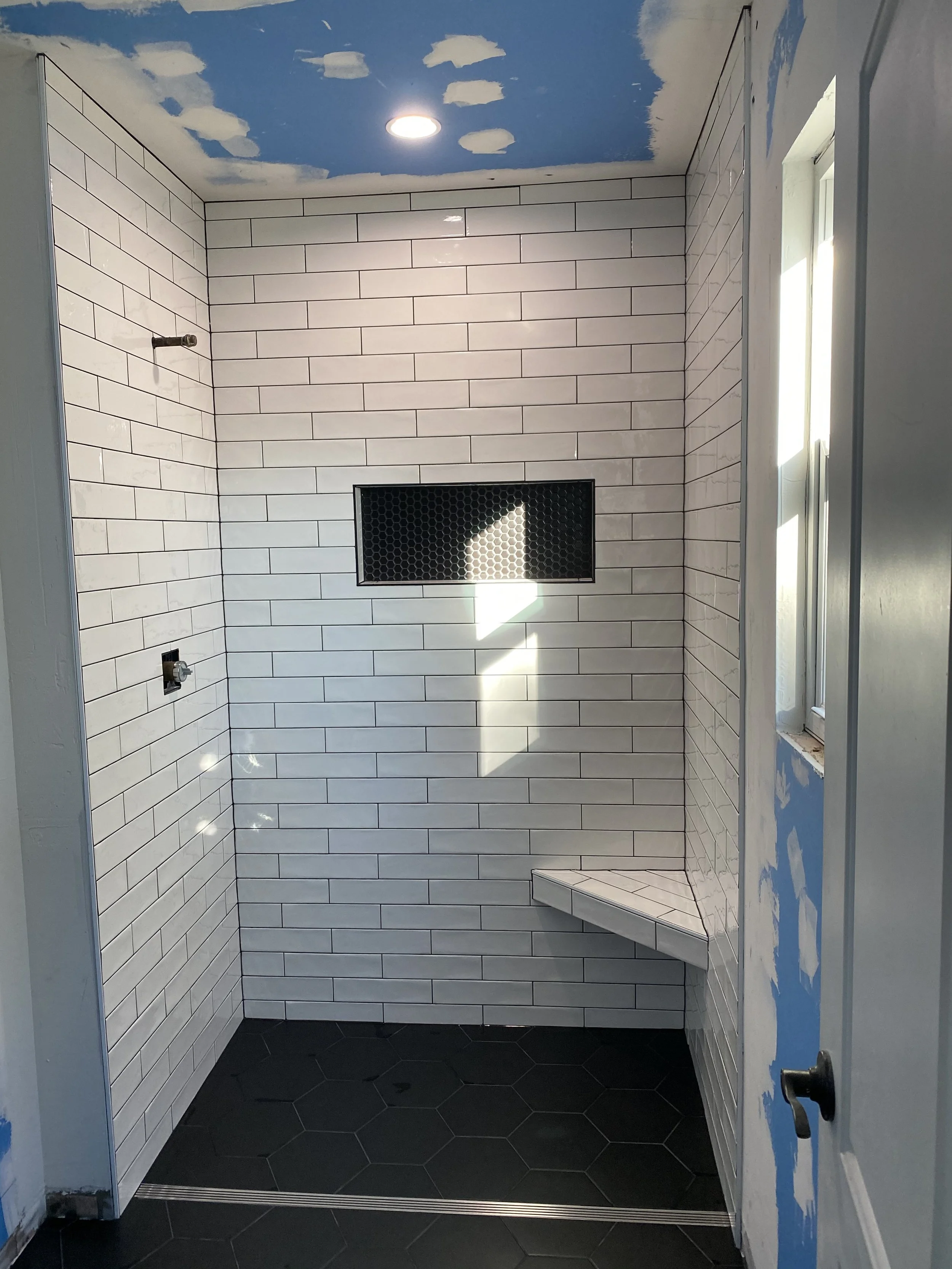 Unfinished bathroom with white subway tile walls, black hexagon tile floor, and a blue ceiling decorated with white clouds. There is a window on the right wall, a small built-in corner bench, and black fixture holes for a shower or faucet.