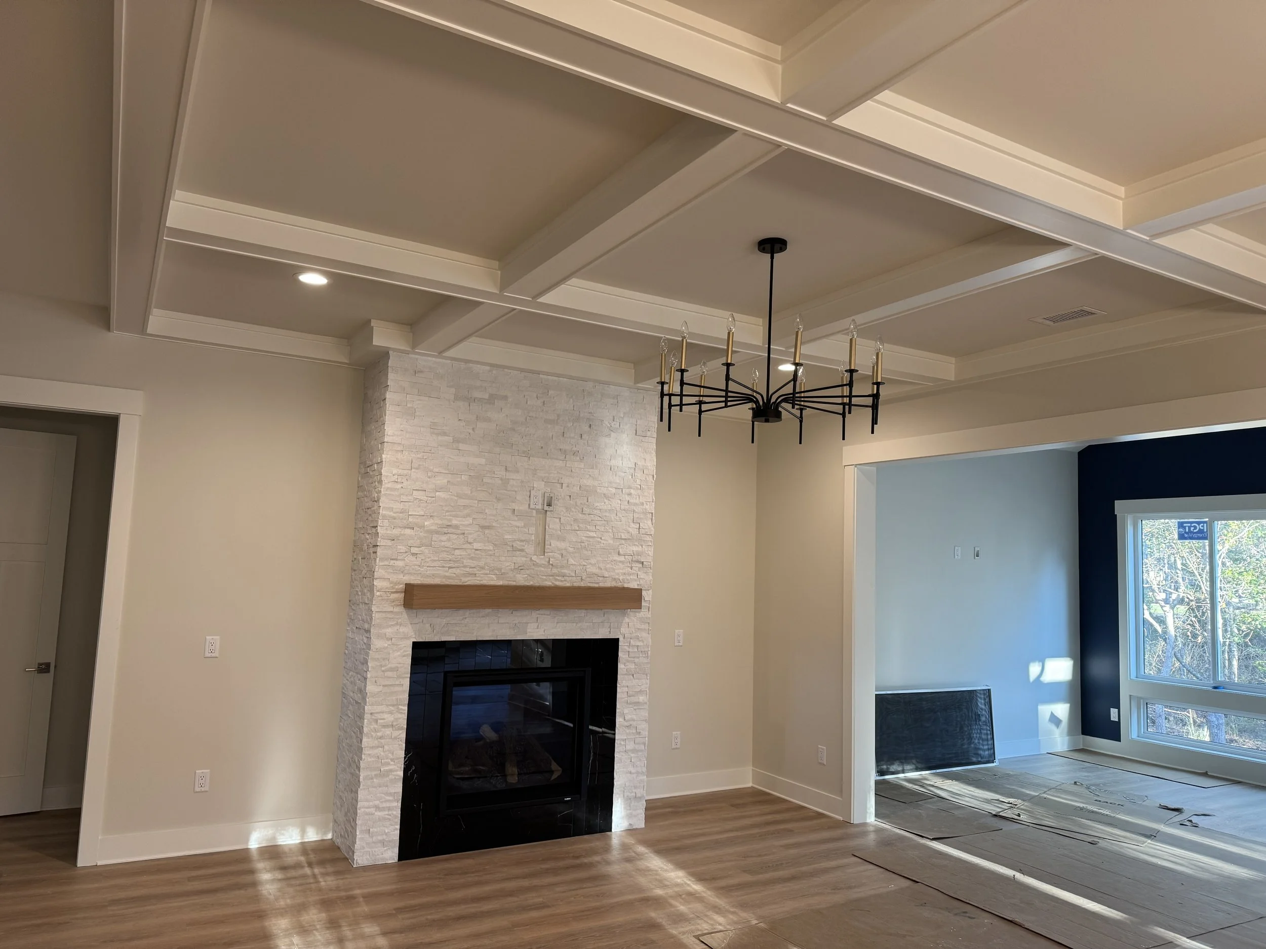 Empty living room with a white stone fireplace, black framed glass door, wood floor, and a black chandelier hanging from the ceiling. There is an opening leading to a room with a blue accent wall, large window, and some construction materials on the 