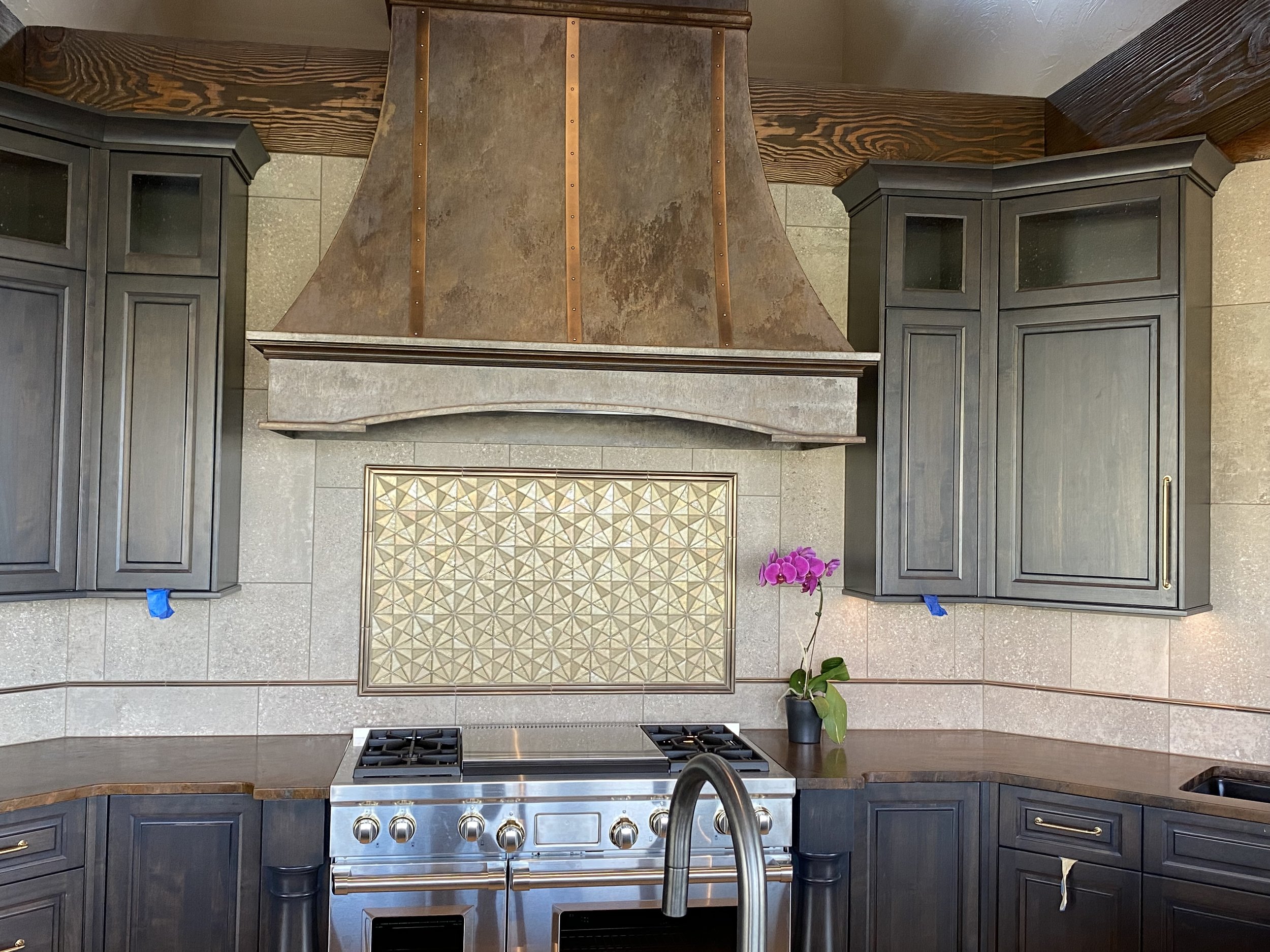 Kitchen with dark cabinets, a stovetop with oven, a copper range hood, a decorative backsplash, a purple orchid plant on the counter, and a sink.