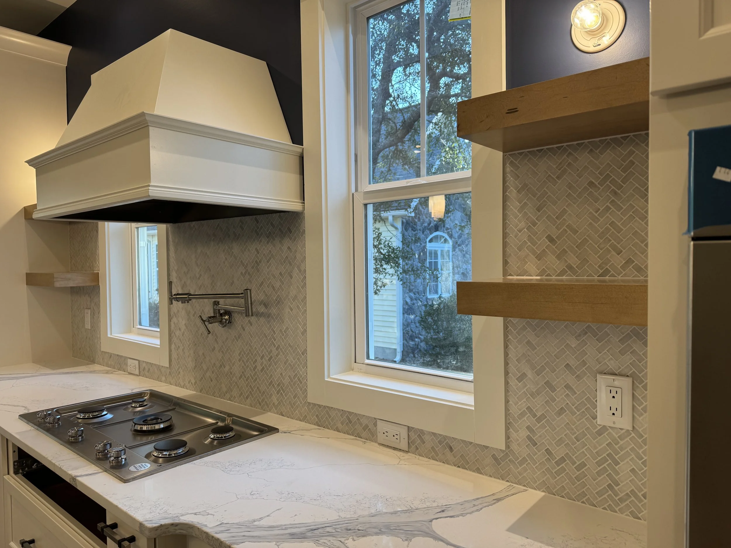 Kitchen with marble countertop, three gas burners, and a backsplash with small herringbone tiles. Two windows above the counter reveal trees outside. There are floating wooden shelves on the wall and a range hood overhead.
