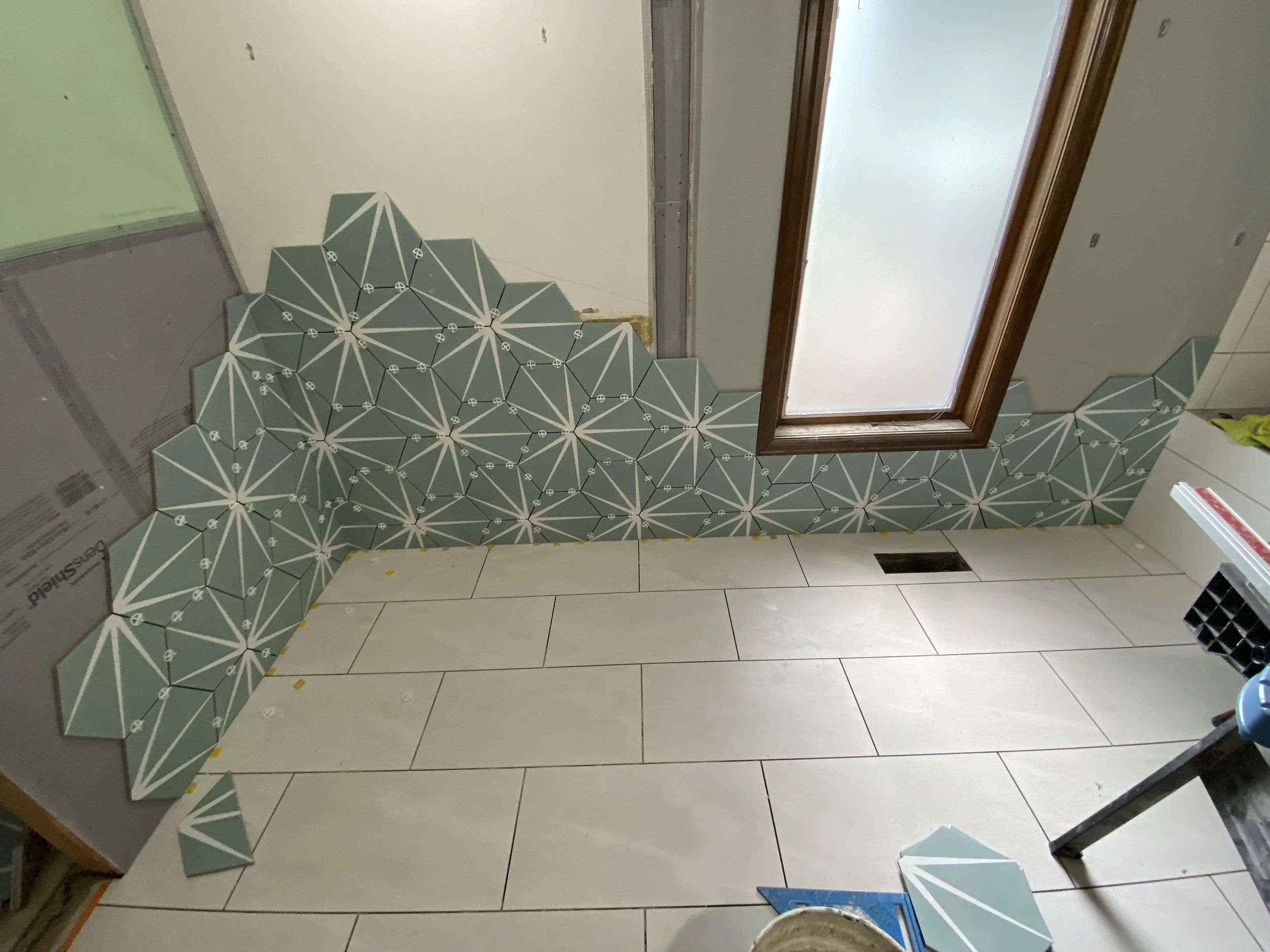 Interior room under construction with partially tiled walls displaying geometric patterned tiles around a window opening, and tiled flooring with some tools and materials visible.