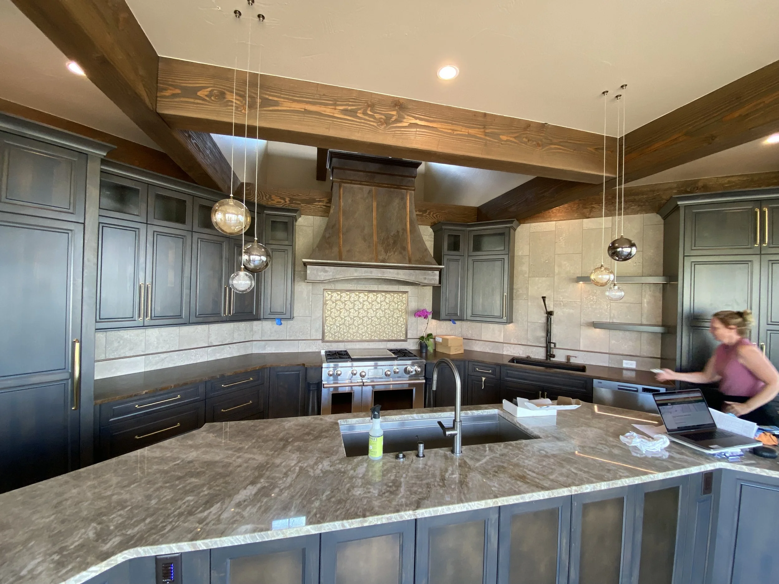 Modern kitchen with gray cabinets, a large marble island, a stainless steel stove with a copper range hood, pendant lights, and a woman in a pink top moving near the right side of the counter.