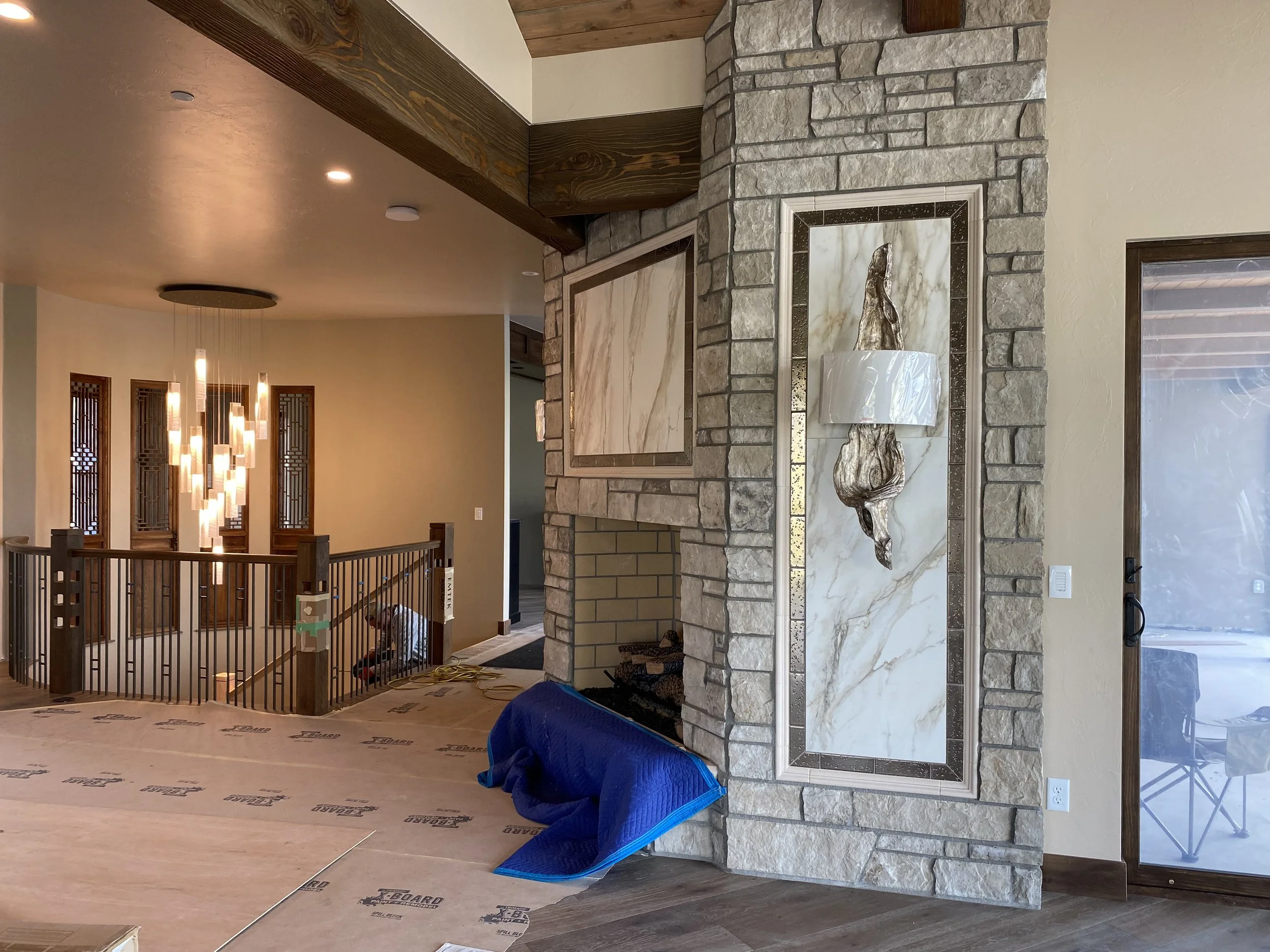 Interior of a home under construction with a fireplace, stone and marble wall, and staircase. A blue protective covering is on the floor. There is a sliding glass door on the right, and a modern chandelier hanging above the staircase.