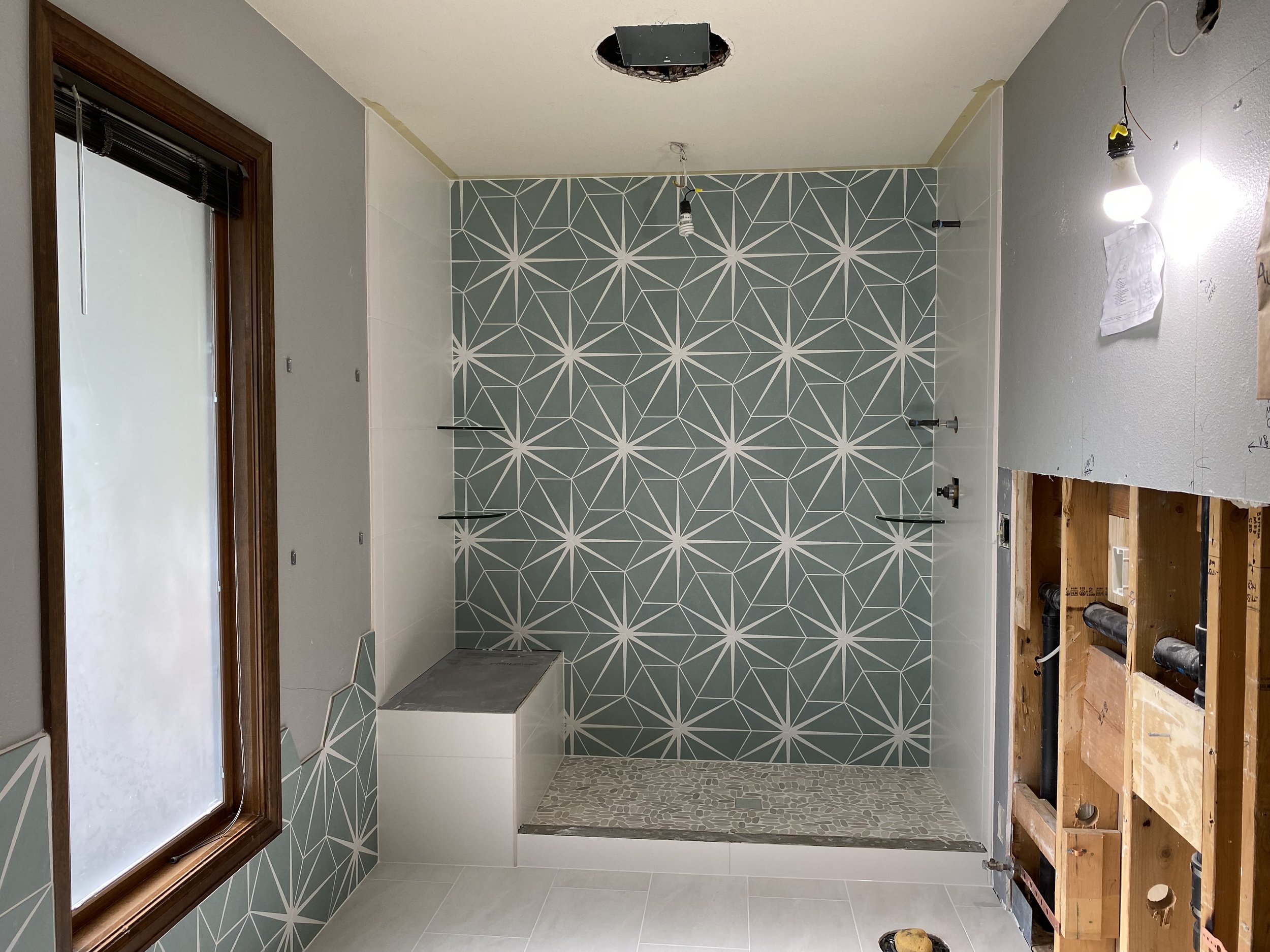 Bathroom under renovation with partially tiled shower area, geometric patterned tiles on the wall, a window with wooden frame, an unfinished area with exposed plumbing and wall, and temporary lighting.