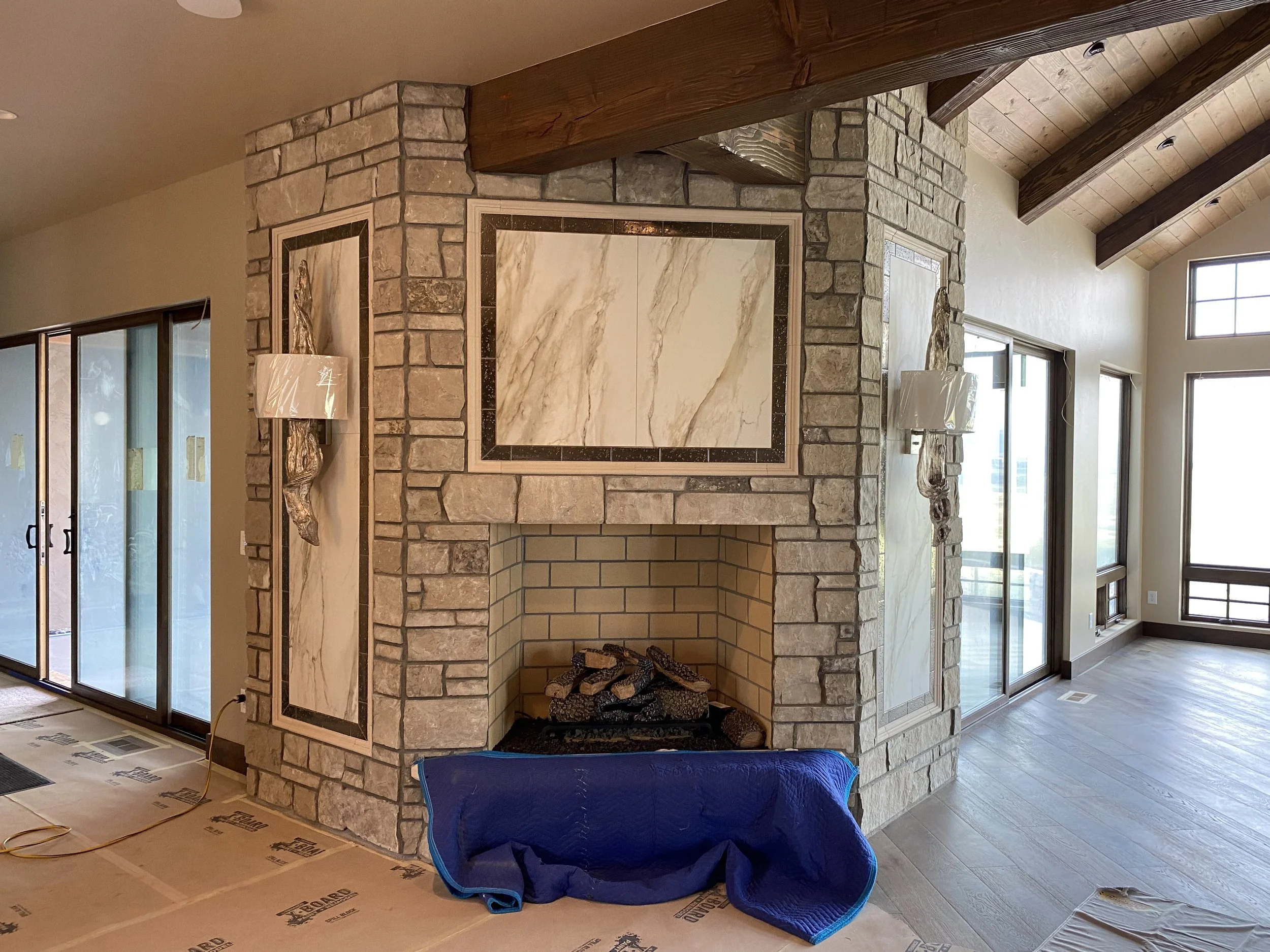 Living room with a stone fireplace, wooden ceiling beams, large windows, and sliding glass doors, currently under renovation with floor covering and construction materials visible.