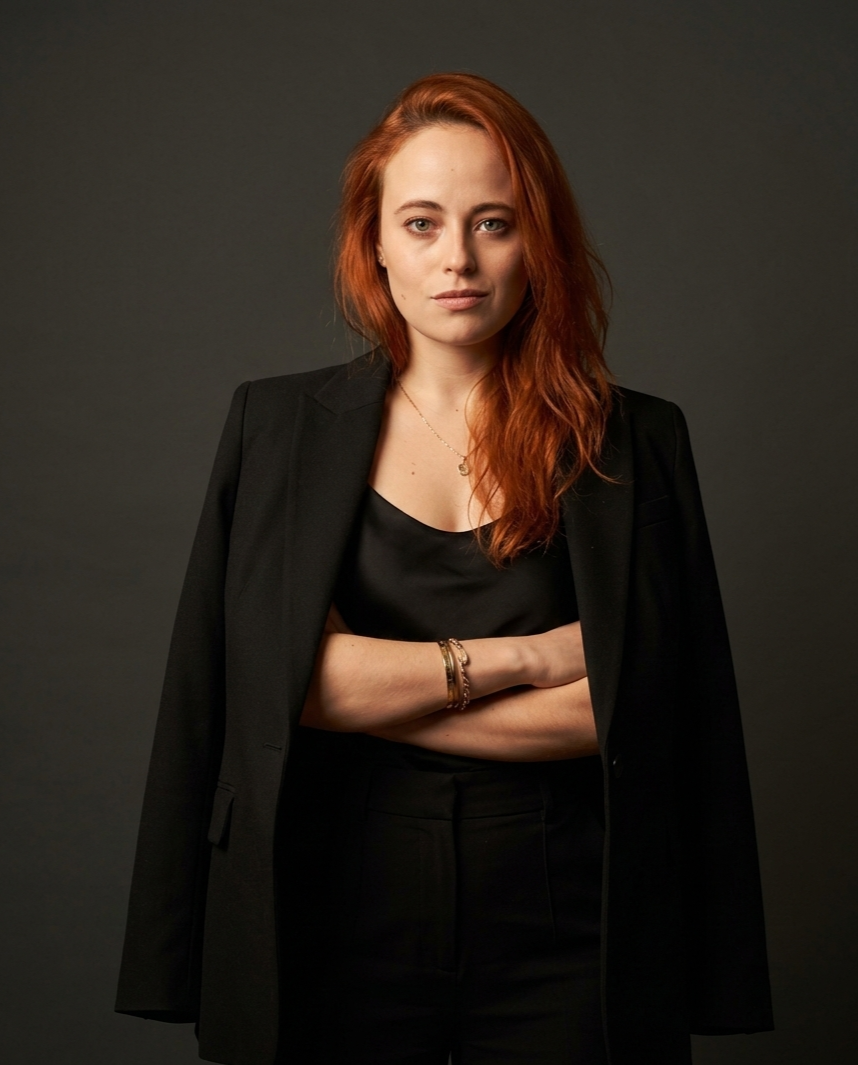 A woman with shoulder-length red hair, dressed in a black blazer over a black top, standing with her arms crossed against a dark grey background.
