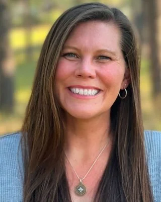 A woman with long brown hair smiling outdoors with a blurred background of trees and yellow flowers.