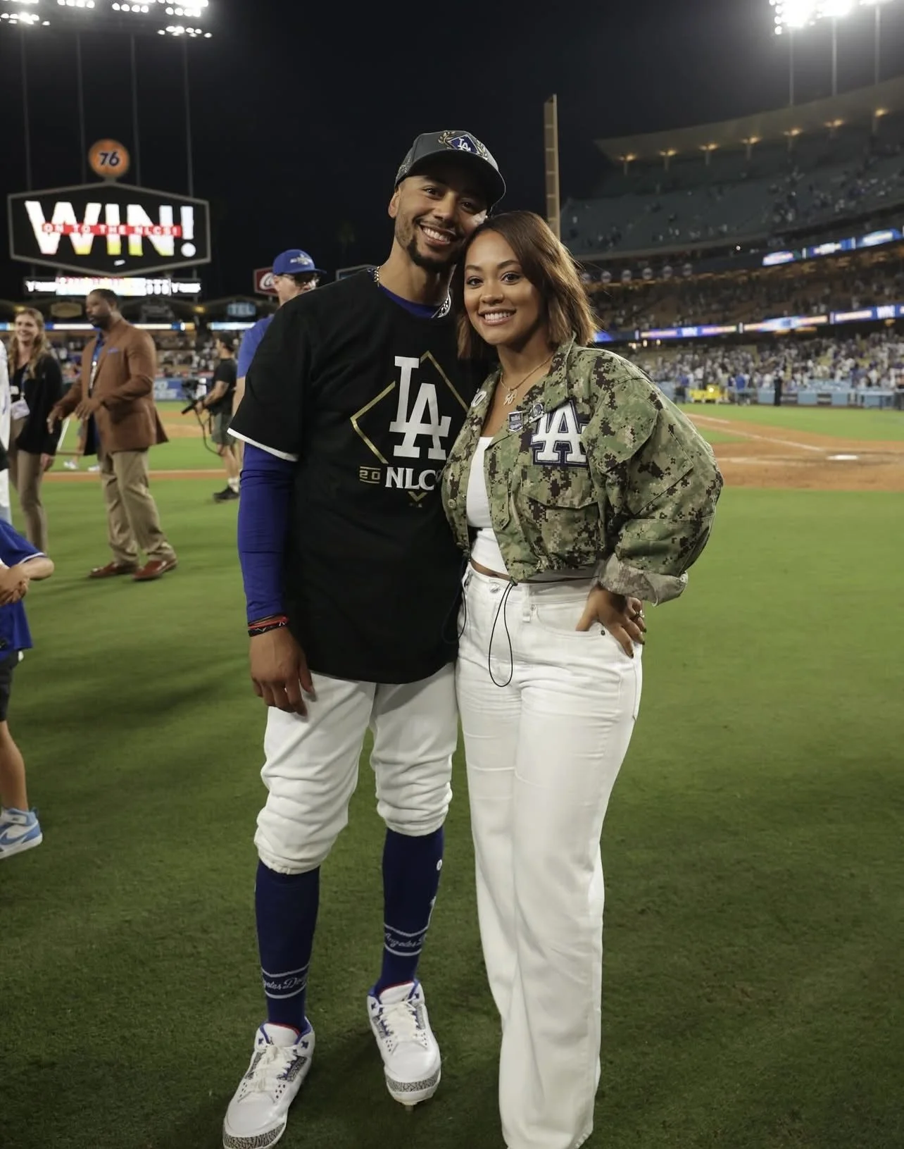 A man in LA Dodgers baseball uniform and a woman in a camouflage jacket pose together on a baseball field at night, smiling for the camera. Several people and a large scoreboard with 'WIN!' are visible in the background.