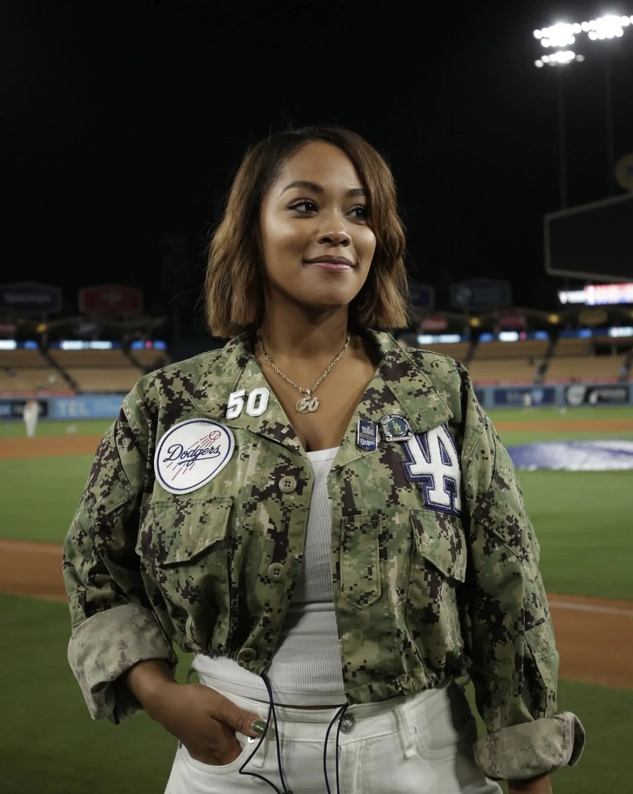 A woman in military camouflage jacket standing on a baseball field at night, with stadium lights overhead, wearing a Dodgers patch and various pins, and smiling softly.