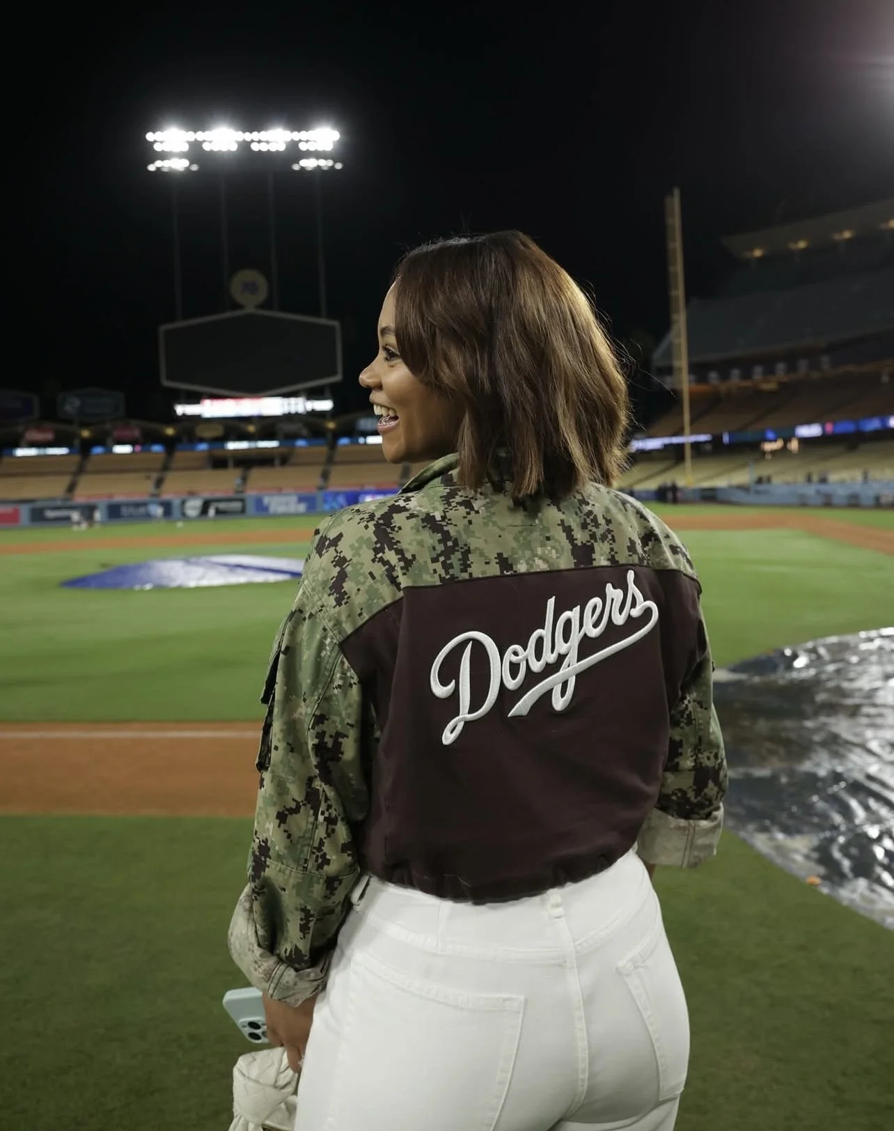 A woman with shoulder-length brown hair wearing a Los Angeles Dodgers jacket and white pants, standing in a baseball stadium at night.