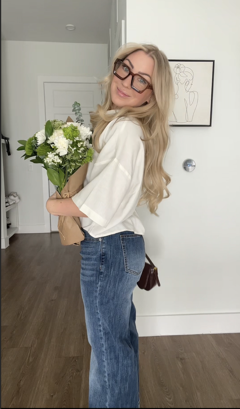 A woman with long blonde hair, wearing glasses, a white top, and blue jeans, is holding a bouquet of white and green flowers while smiling in a modern, minimalist home interior.