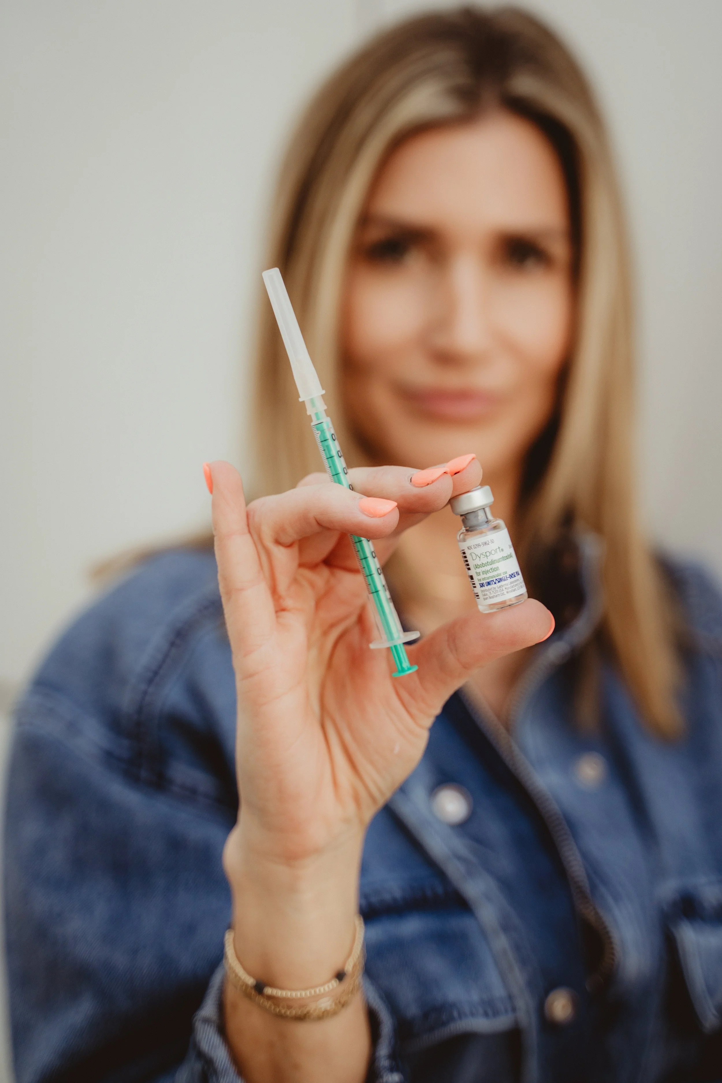 Woman holding a syringe and vaccine vial, smiling, wearing a blue denim jacket, with blond hair, blurred background.