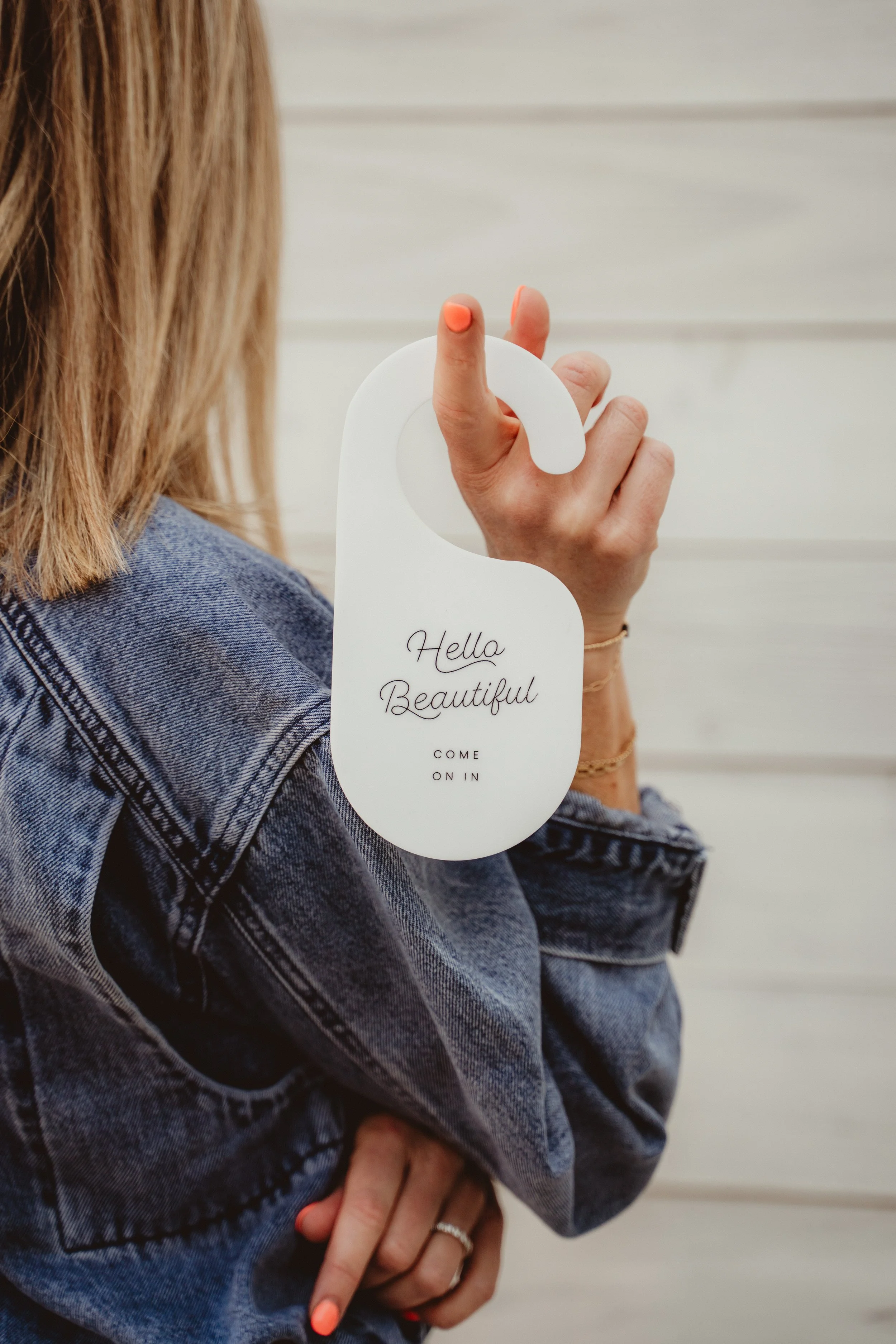 A person holding a white door hanger with the text 'Hello Beautiful Come On In,' wearing a denim jacket and standing in front of a light-colored wooden wall.