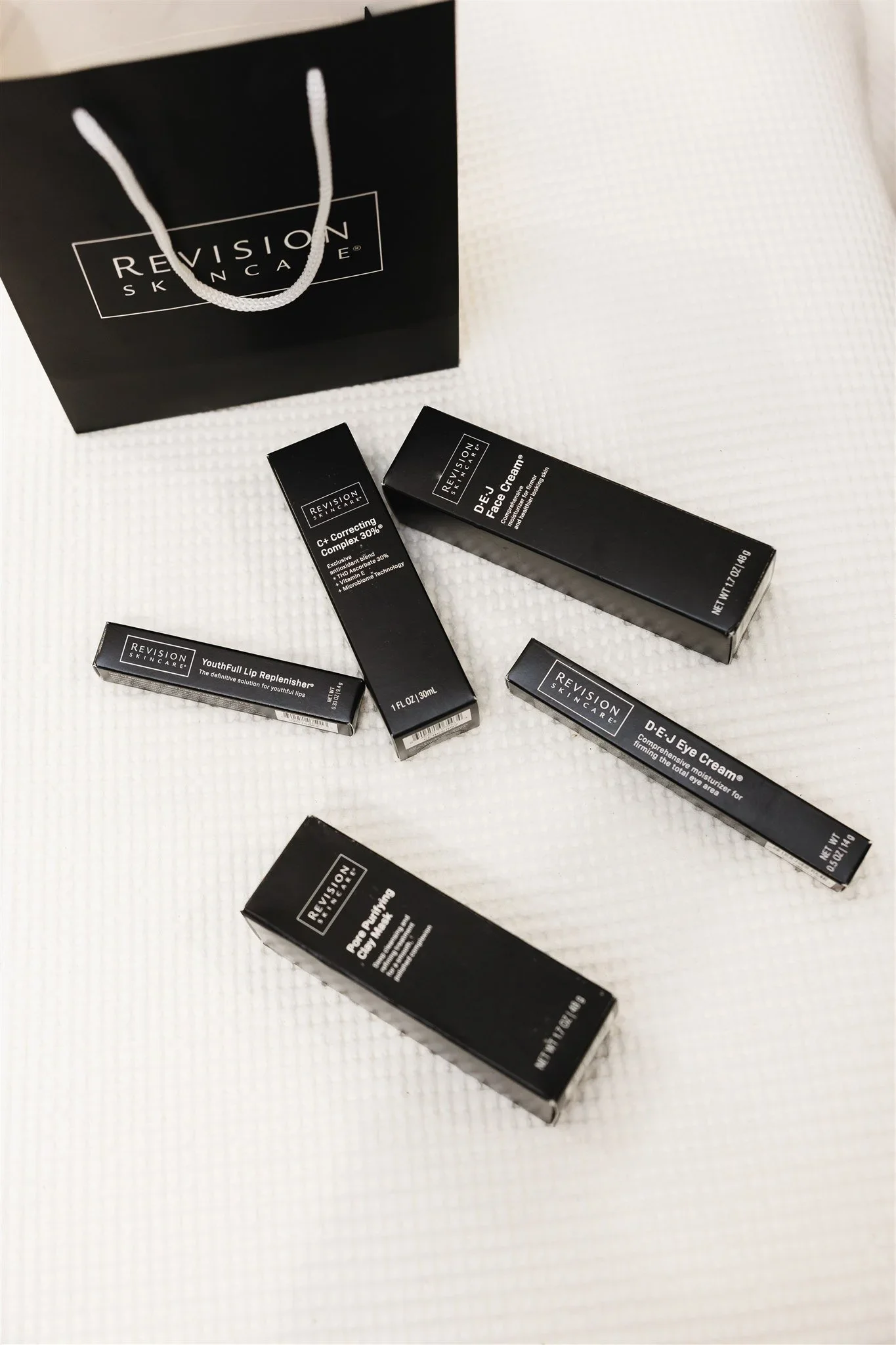 Assorted black and white Revisions Skincare cosmetic products, including creams and lip filler, displayed on a white textured surface with a black shopping bag in the background.