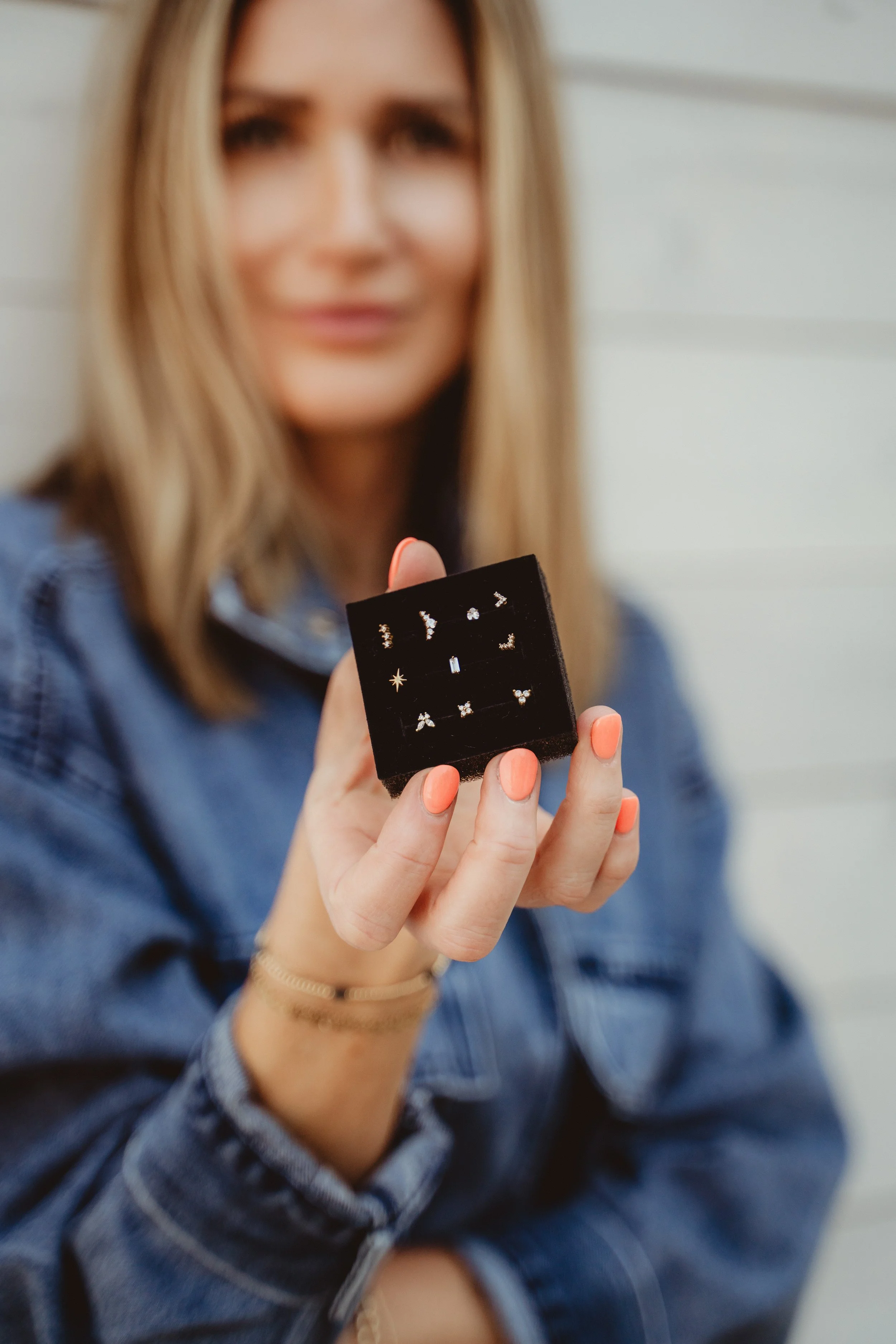 Woman in denim jacket holding a small black jewelry box with earrings, with a white wall background.