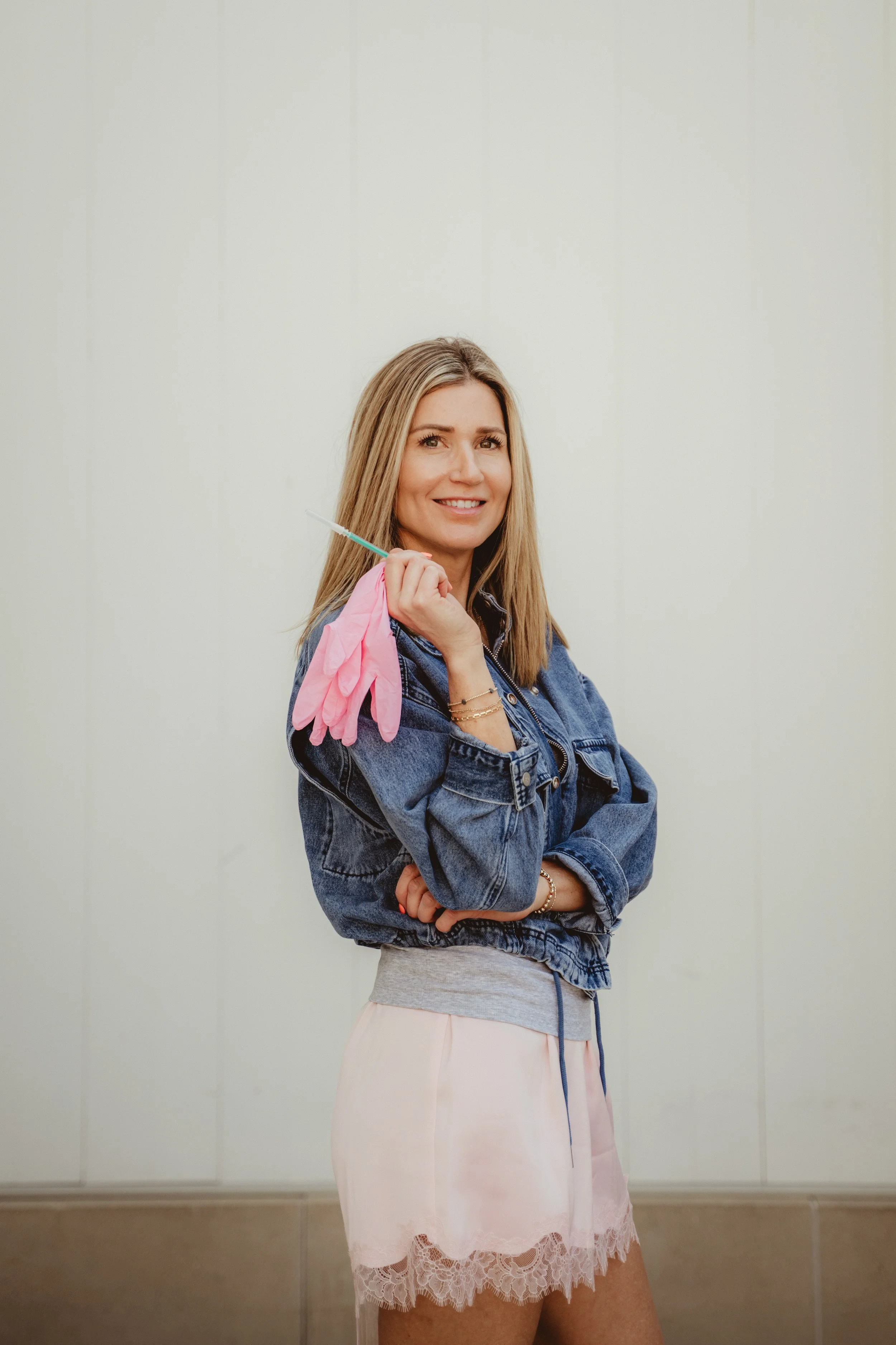 Woman in denim jacket holding pink rubber gloves and syringe, smiling, standing against a plain wall.