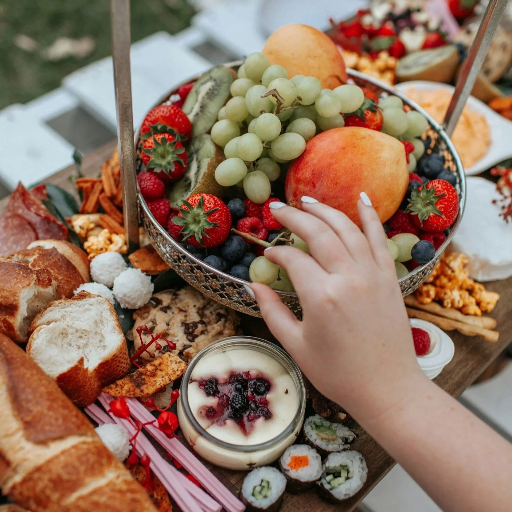 A fruit platter featuring strawberries, green grapes, peaches, and kiwi, with assorted sushi rolls, bread, and a berry dessert on a wooden table.