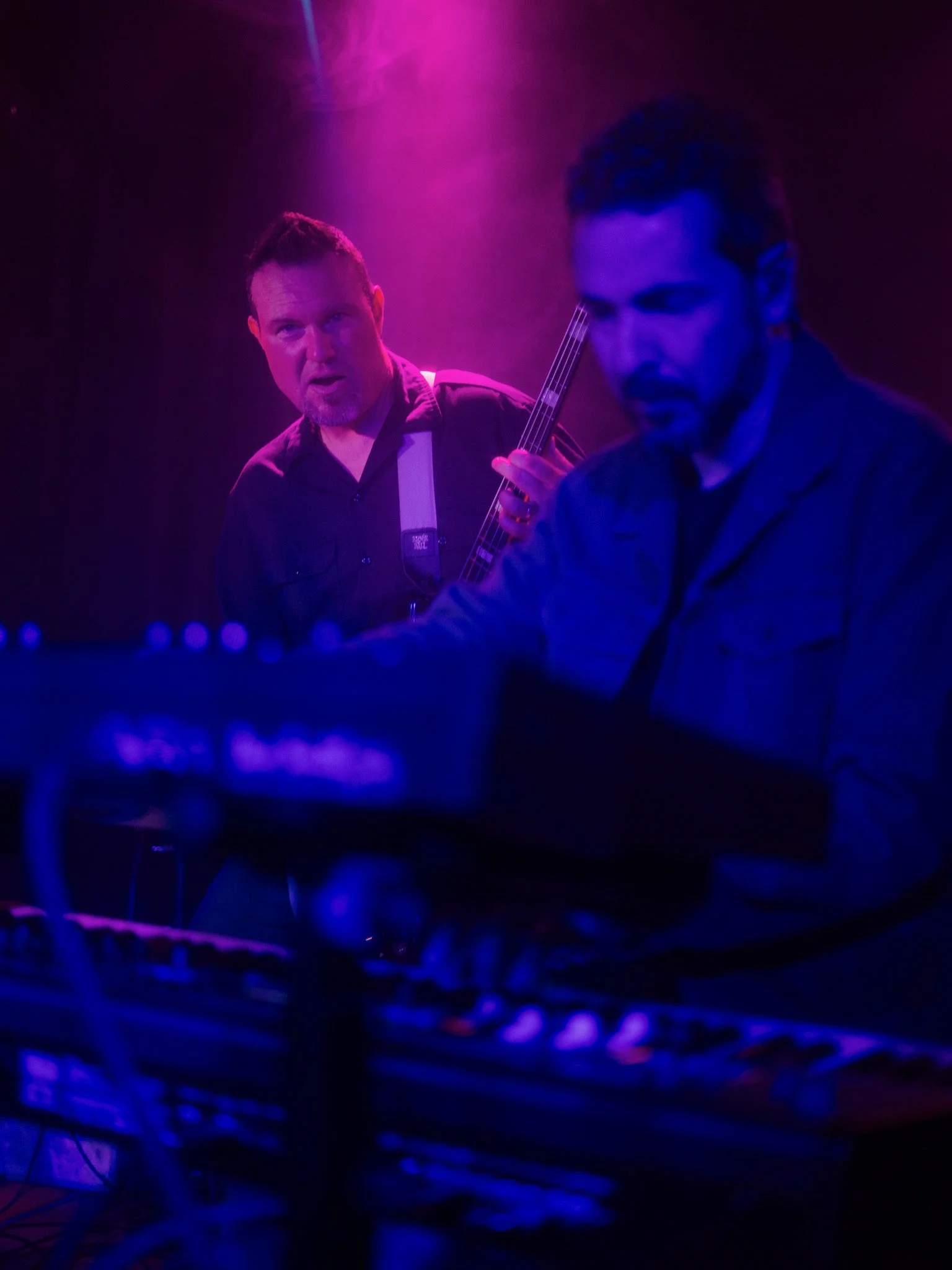Two musicians perform on stage with purple lighting; one plays keyboard, the other holds a bass guitar and looks towards the camera.