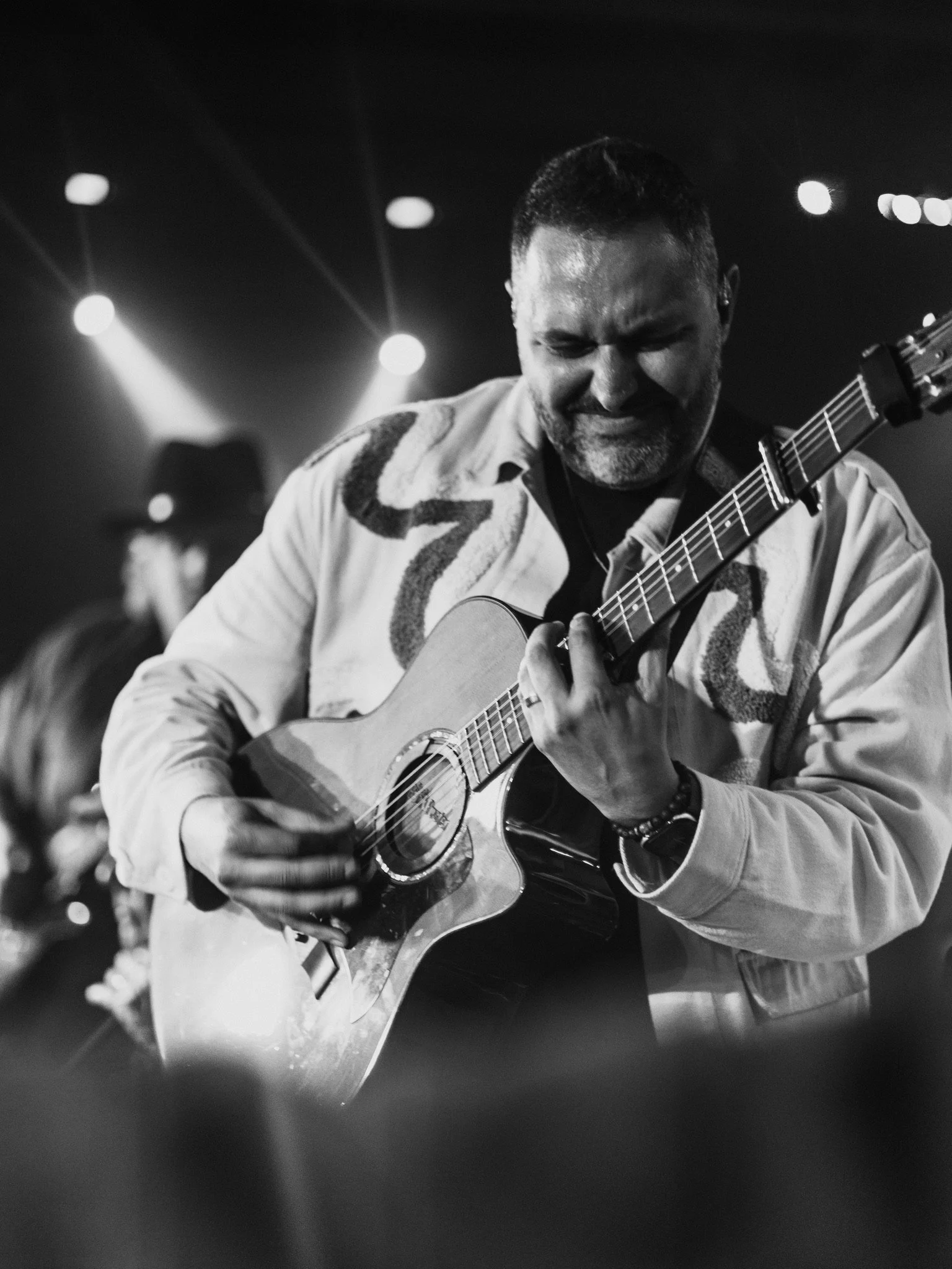 Man playing acoustic guitar on stage with lights in the background, black and white photo.