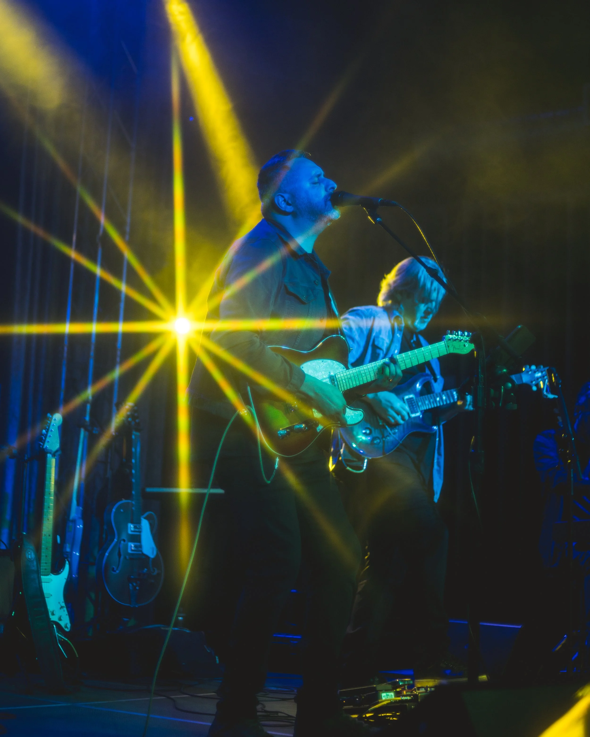 Two musicians playing guitars on stage under bright yellow and blue stage lighting, with guitars and equipment in the background.