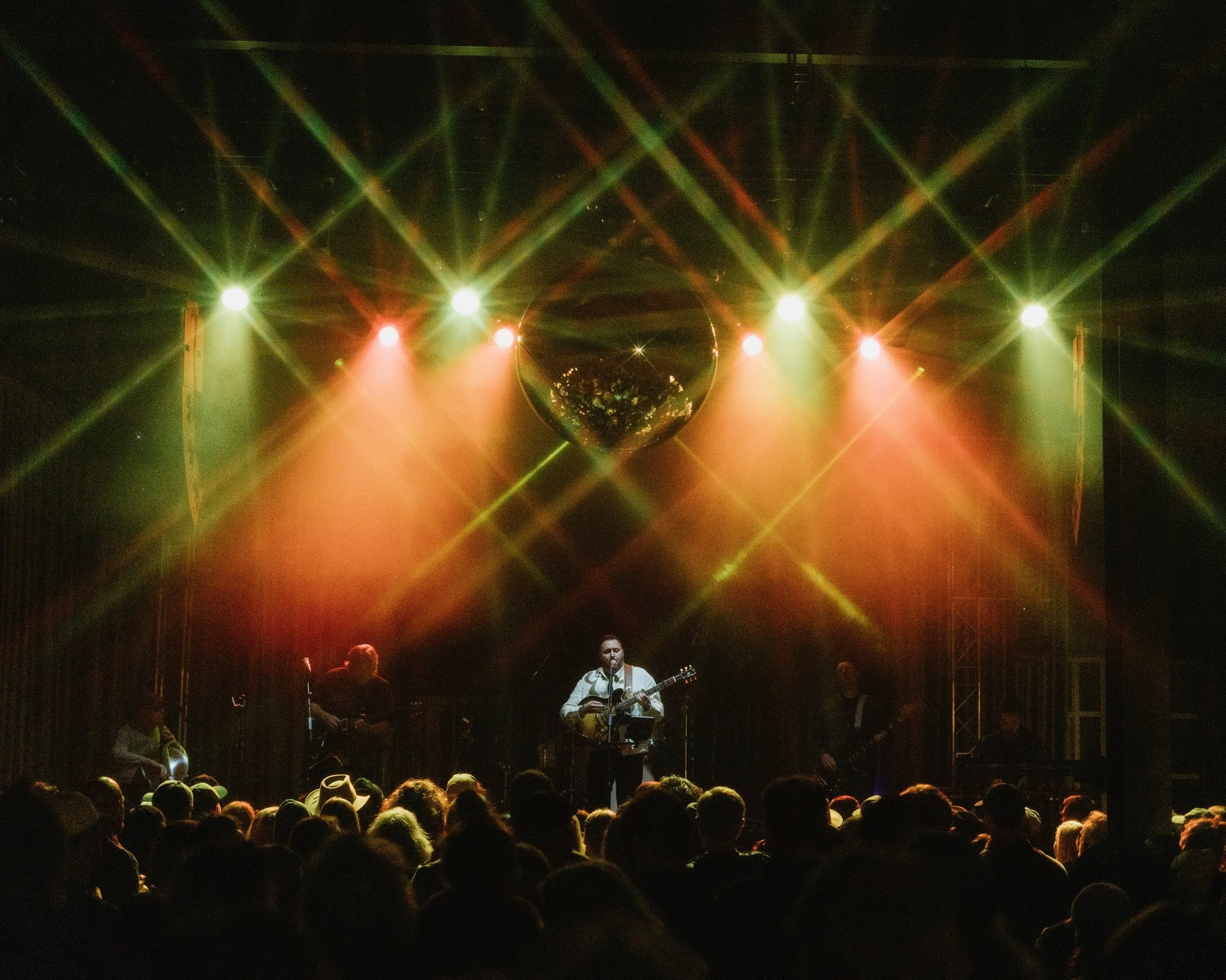 A musician plays guitar and sings on stage during a concert, with bright lights and a disco ball overhead, in front of an audience.