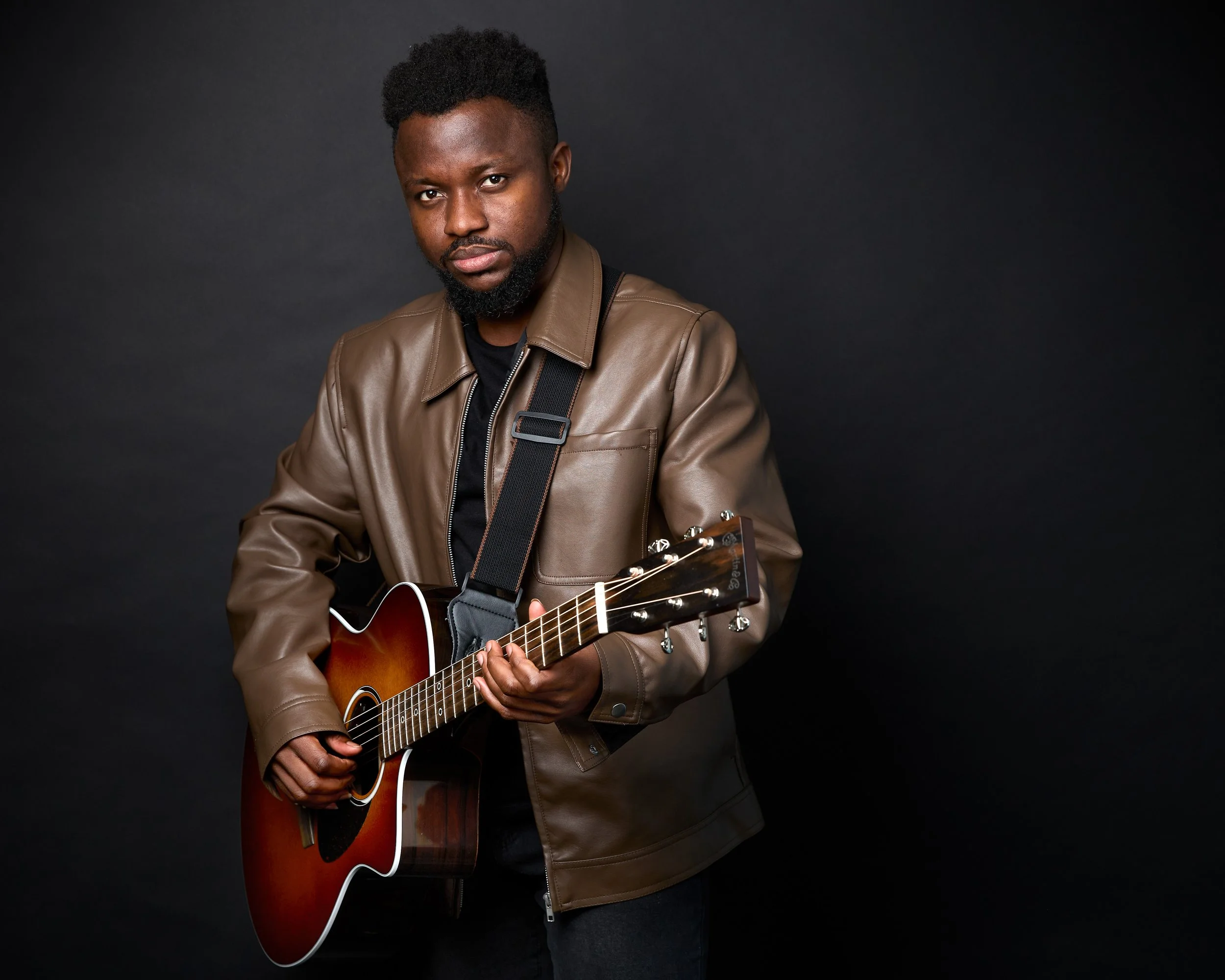 A young man with dark skin and a beard playing an acoustic guitar against a black background, wearing a brown leather jacket and a black shirt.