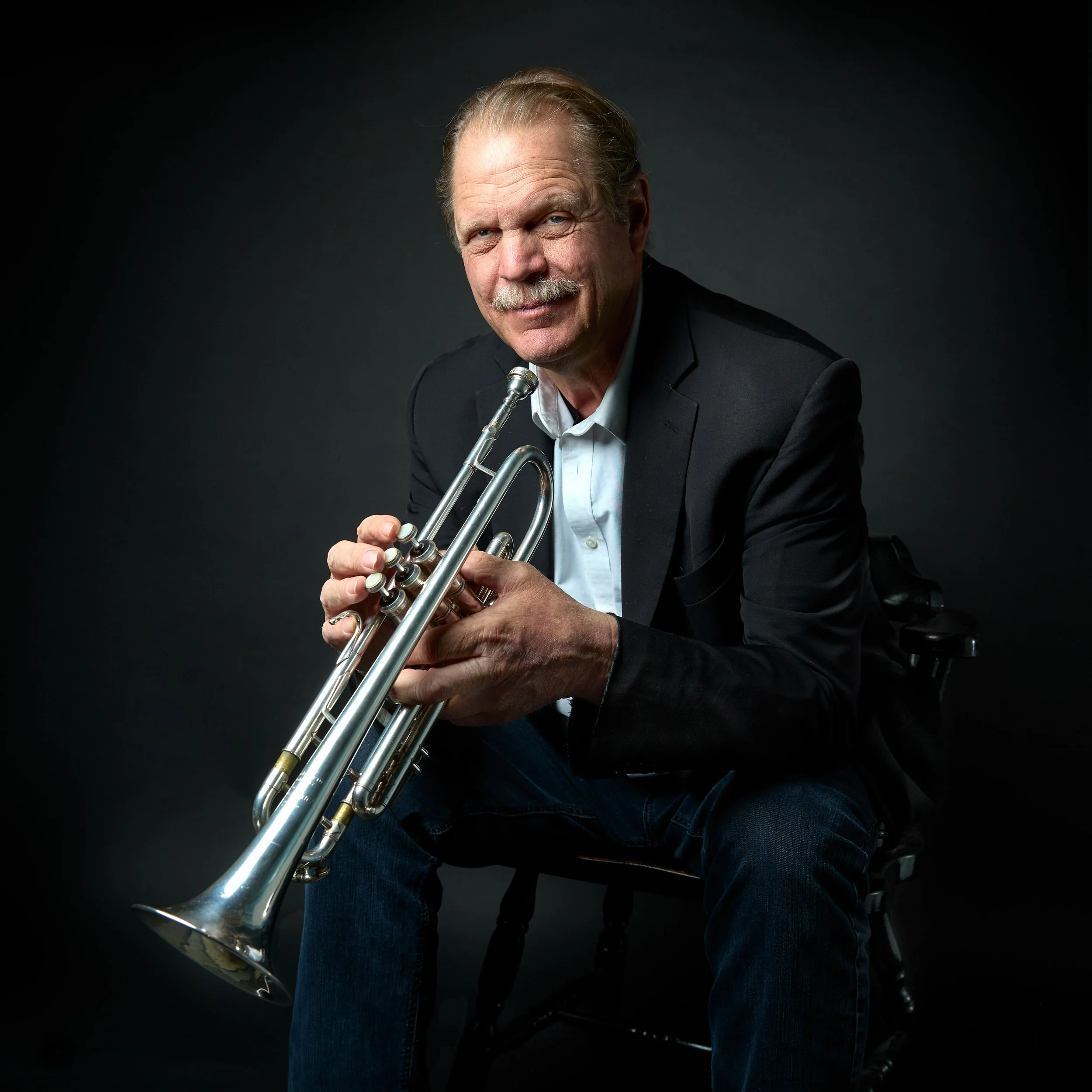 An older man with a mustache wearing a black blazer, white shirt, and jeans sitting on a black chair holding a silver trumpet against a dark background.