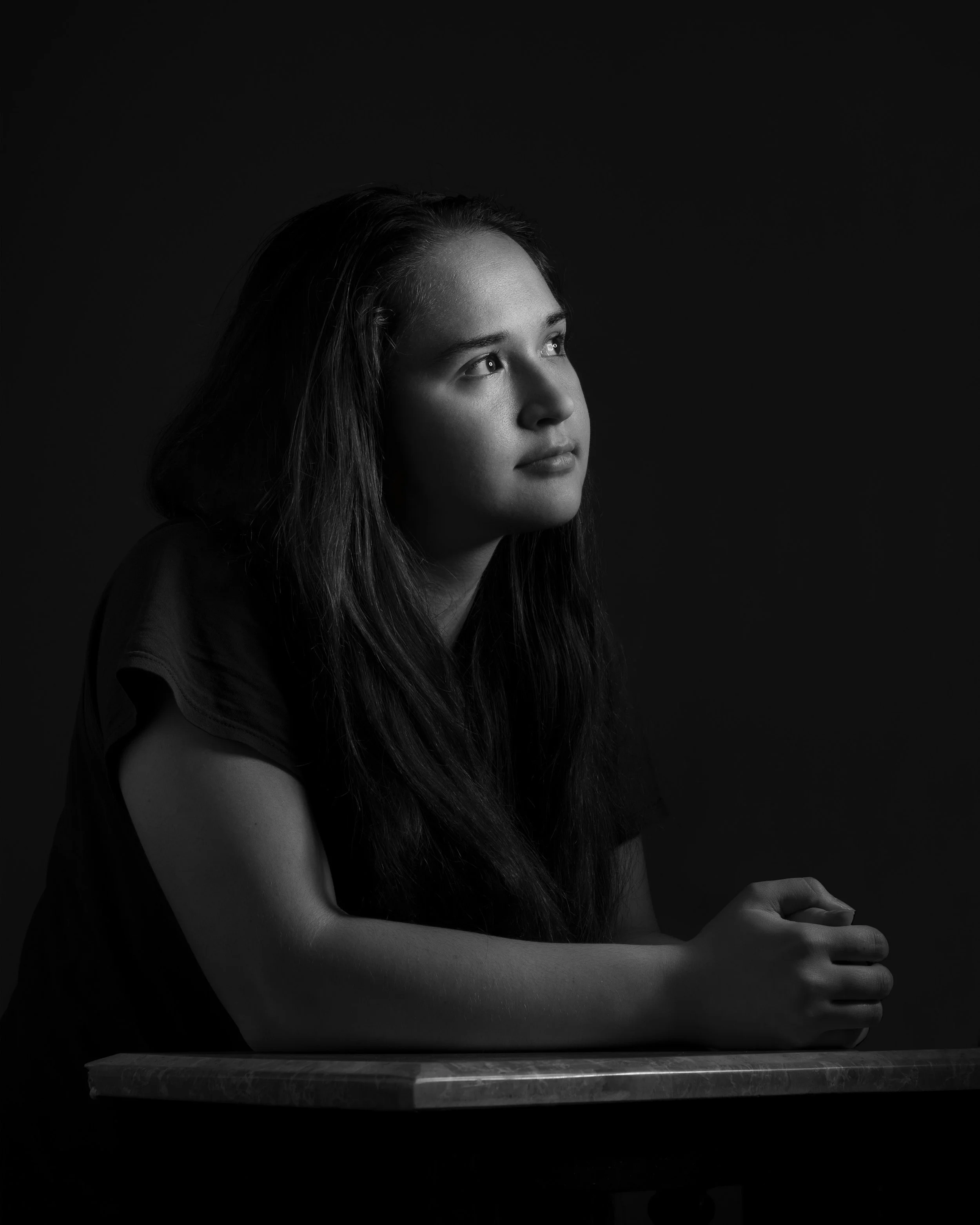 Black and white portrait of a young woman with long hair, resting her arms on a table and looking off to the side with a thoughtful expression.