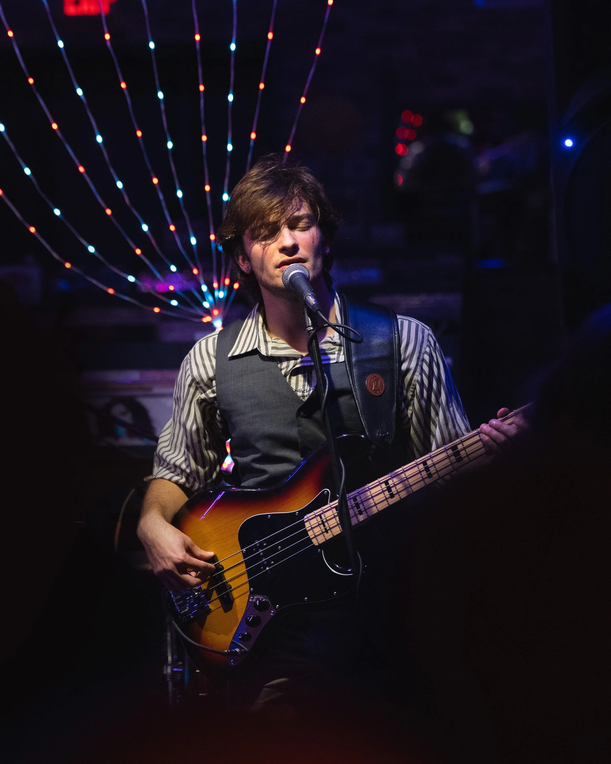 Musician singing into a microphone and playing a bass guitar on stage with colorful stage lights in the background.