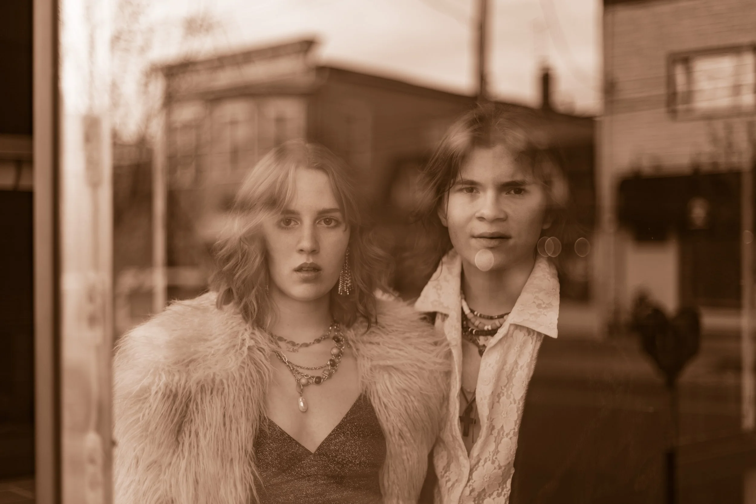 Two young adults, a woman and a man, stand close together inside, looking through a window at the camera. They wear vintage-style clothing and jewelry, with a blurry residential neighborhood in the background.