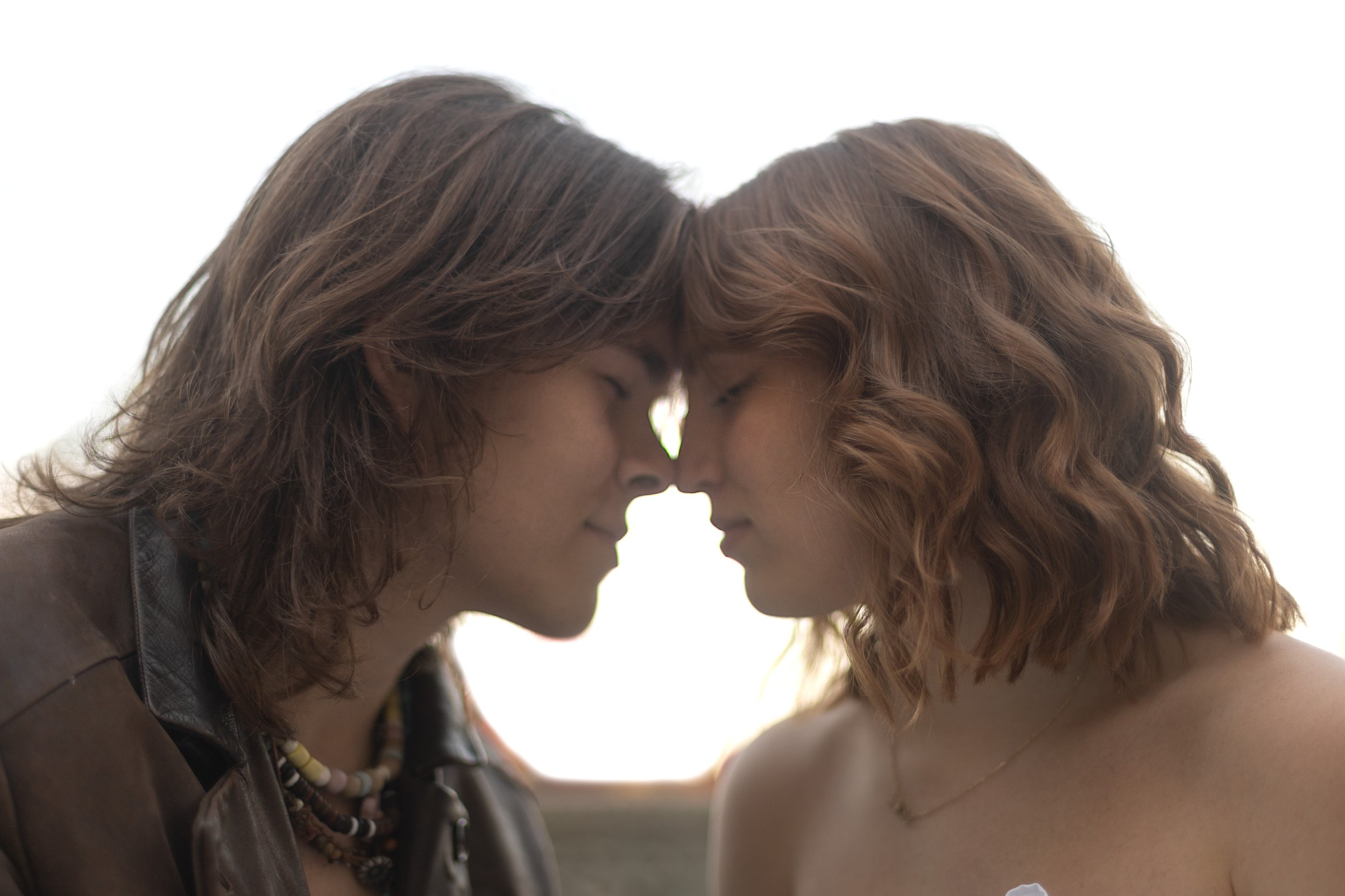 Two women with red hair and light skin touch foreheads and smile with eyes closed, outdoors against a bright sky background.