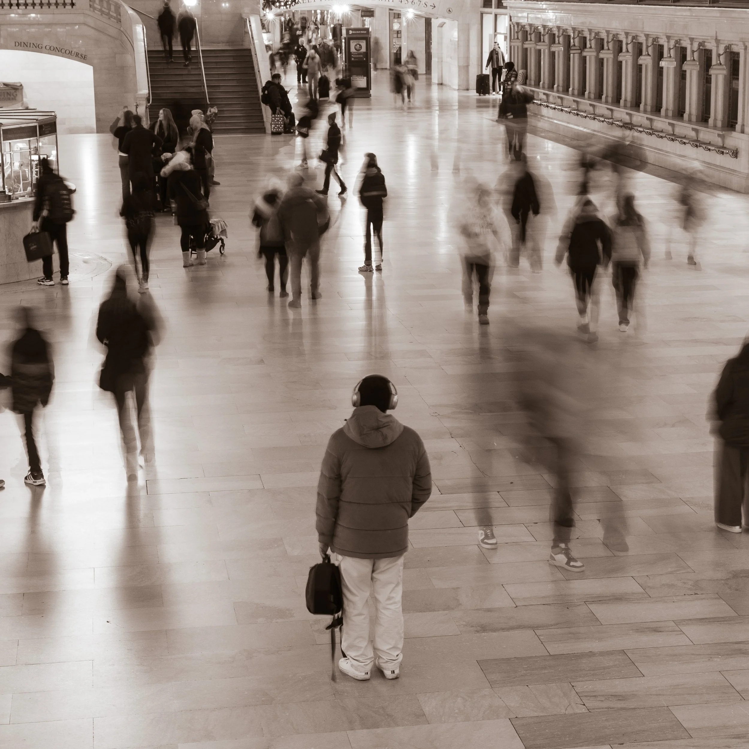 A sepia-toned photo of a busy train station or airport terminal with many blurred pedestrians walking in various directions. In the foreground, a man wearing a hoodie, headphones, and white pants stands alone, holding a bag and looking down.