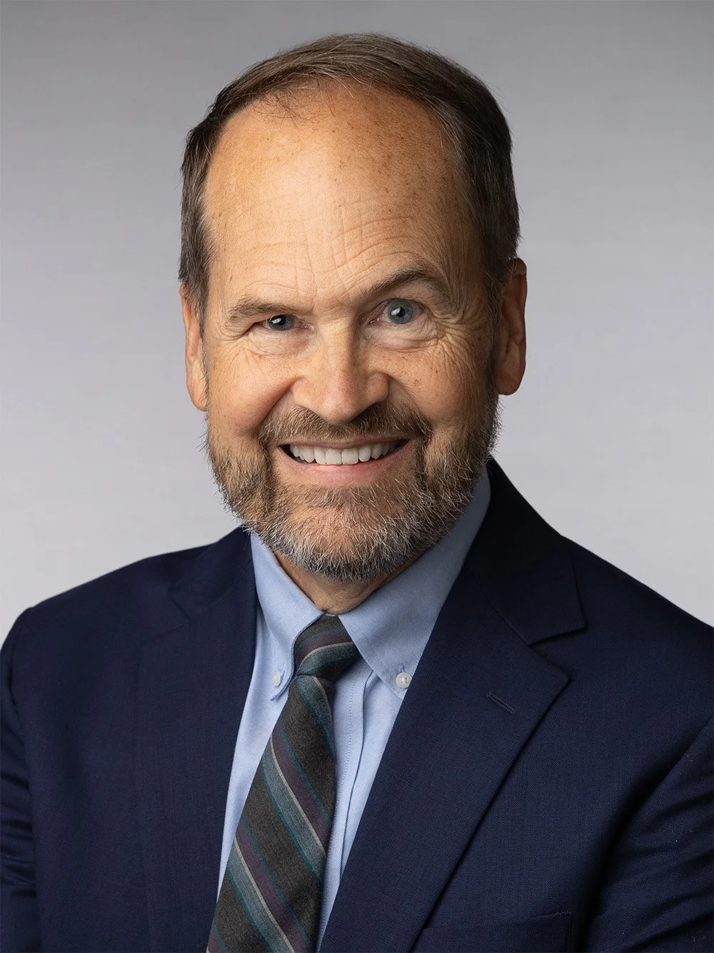 A smiling middle-aged man with a beard and blue eyes, dressed in a dark suit, light blue shirt, and striped tie, posing against a plain gray background.