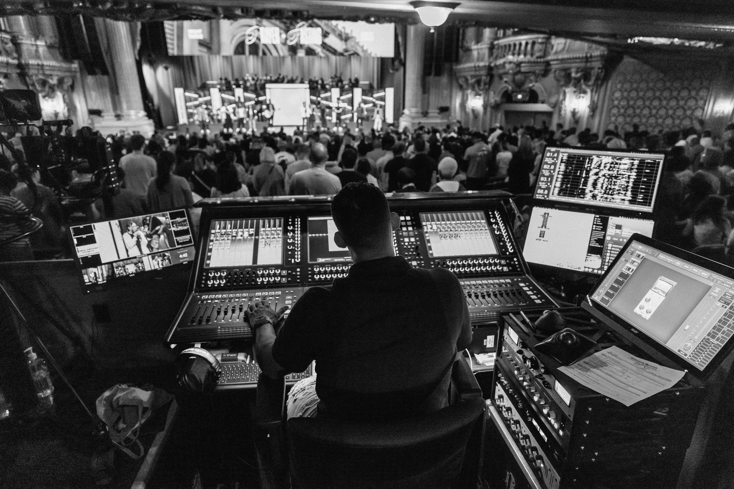 A DJ performing on a stage with a large crowd in a concert hall, seen from behind the DJ's mixing console with multiple screens and audio equipment in black and white.