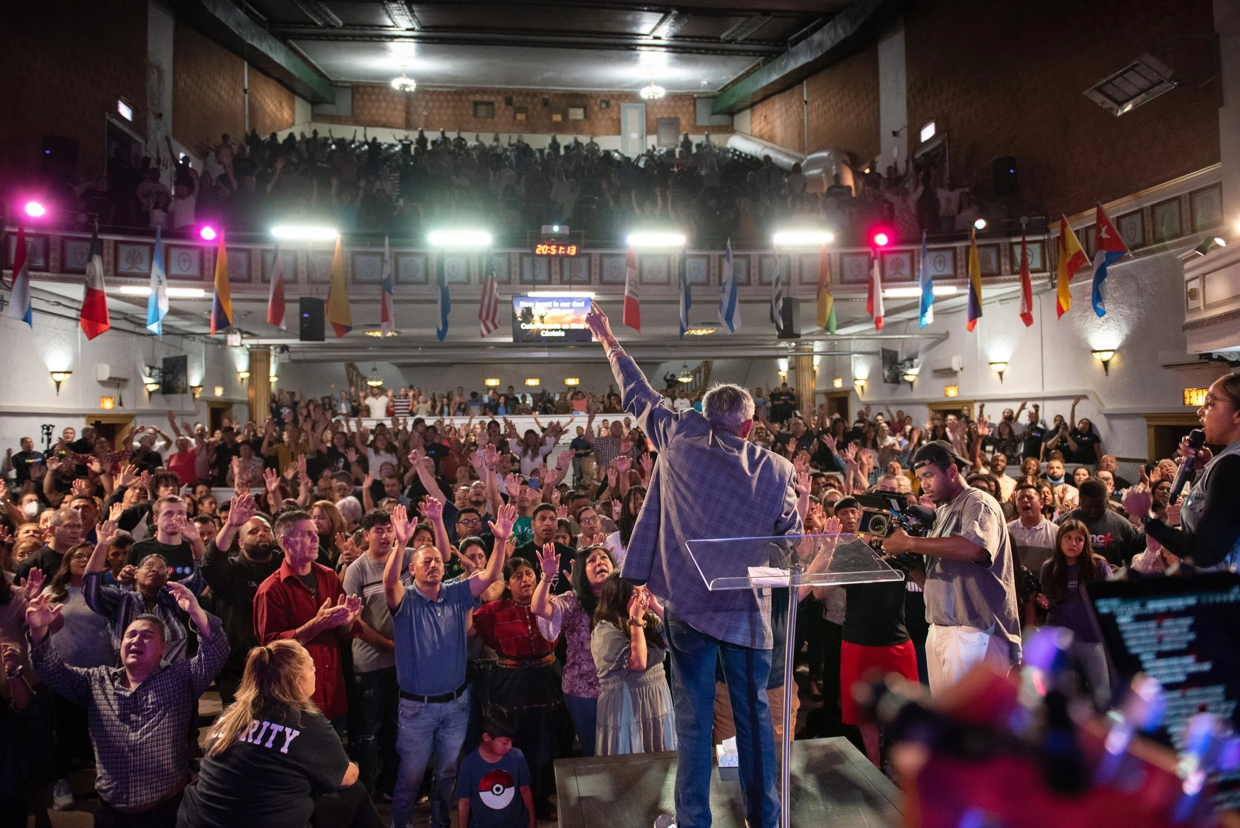 A large indoor gathering with a speaker on stage addressing a crowd. The crowd has many people raising their hands in response. Flags from various countries hang from the ceiling, and there are stage lights and a large screen in the background.