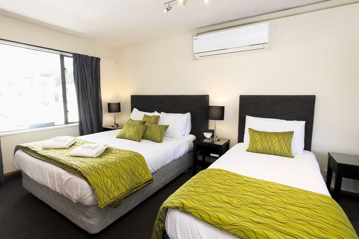 A hotel room with two beds, each with green patterned bedspreads and pillows, black headboards, a nightstand with a lamp, and a window with black curtains.