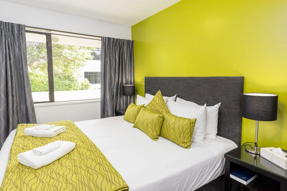 A neatly made bed with a gray headboard, white sheets, and a yellow bed runner with matching pillows. There are two towels on the bed. On either side of the bed, there are black nightstands with black lamps. A window with gray curtains allows natural light into the room, revealing green trees outside. The wall behind the bed is painted bright yellow.
