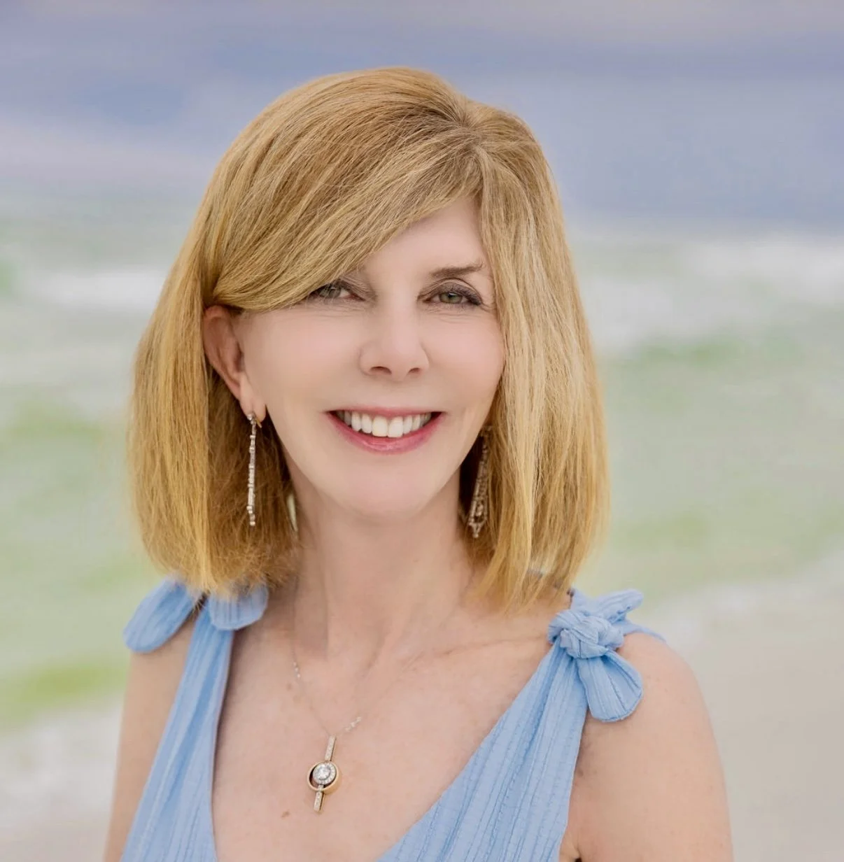 A woman with shoulder-length red hair smiling at the camera, wearing a light blue dress with shoulder bows, earrings, and a necklace, at the beach with the ocean in the background.
