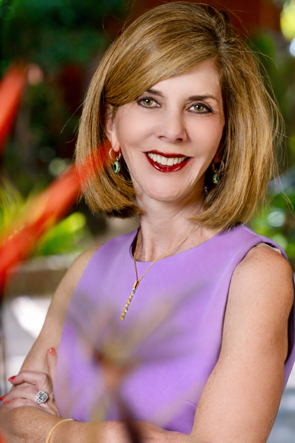 A woman with shoulder-length light brown hair and green eyes, smiling, wearing a purple sleeveless dress, turquoise earrings, a gold necklace with a pendant, and a ring on her right hand. She is standing outdoors with greenery in the background.