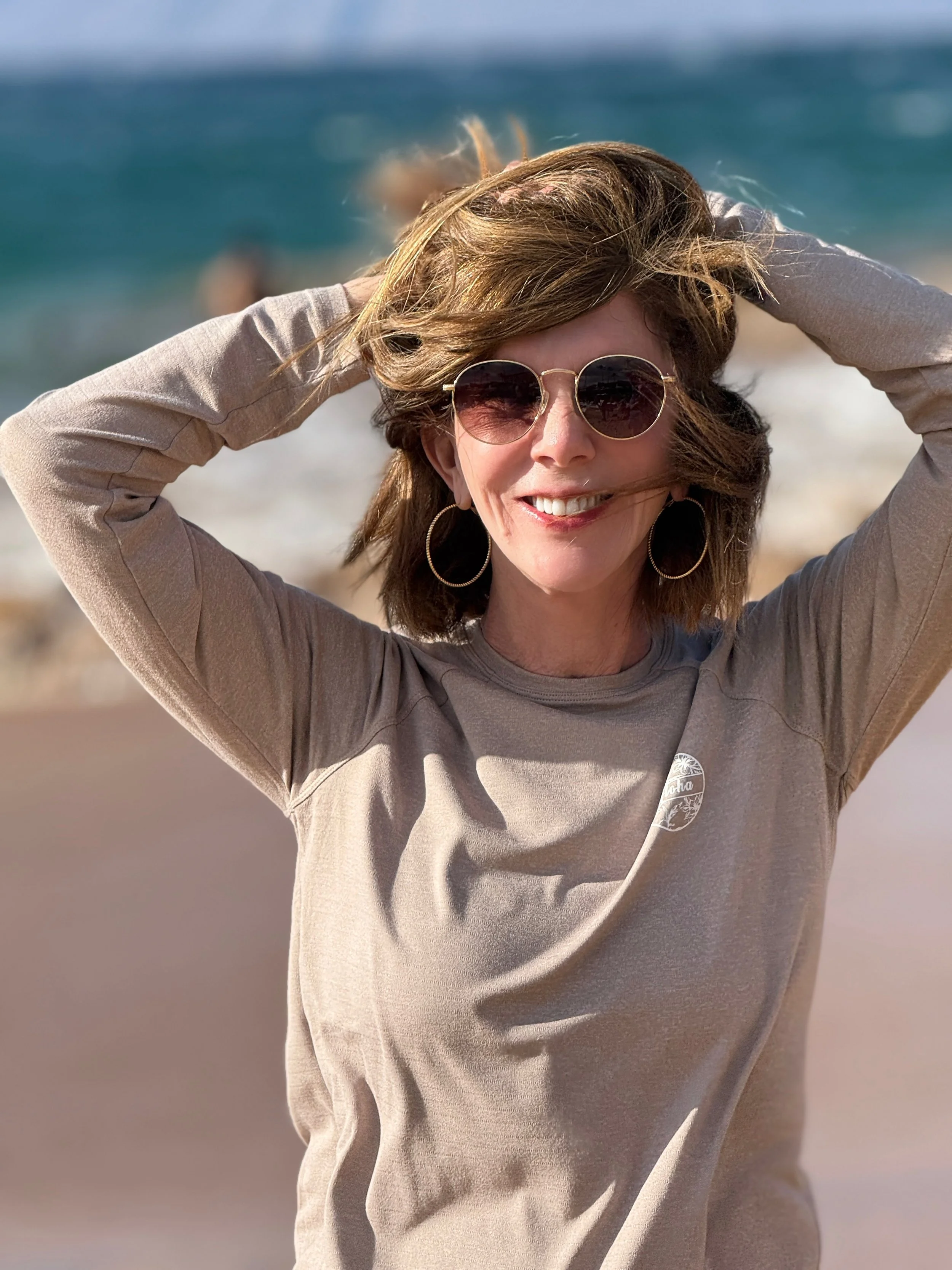 Woman smiling on a beach with sunglasses, earrings, and beige sweatshirt, with ocean and sky in the background.