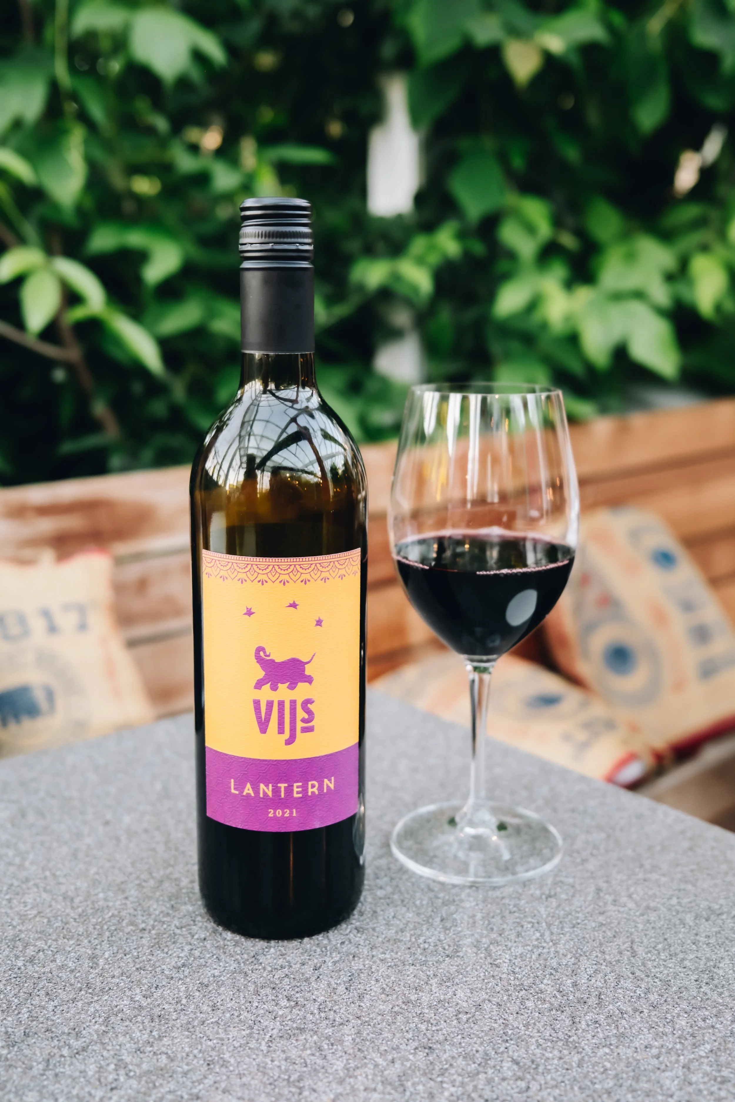 A bottle of dark red wine labeled 'VIJS Lantern 2021' alongside a glass partially filled with red wine on a grey table, with a green leafy background.