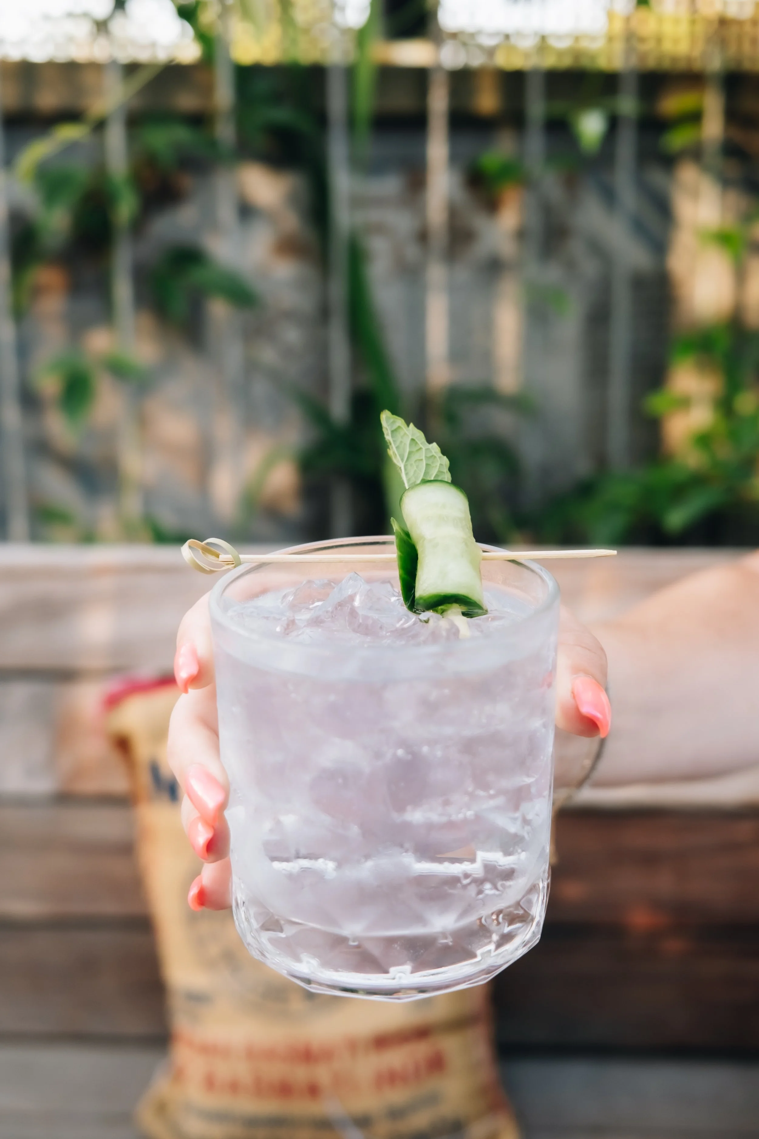 A hand holding a clear glass filled with ice and a light pink drink, garnished with a cucumber slice and a small leaf, with a blurred outdoor garden background.