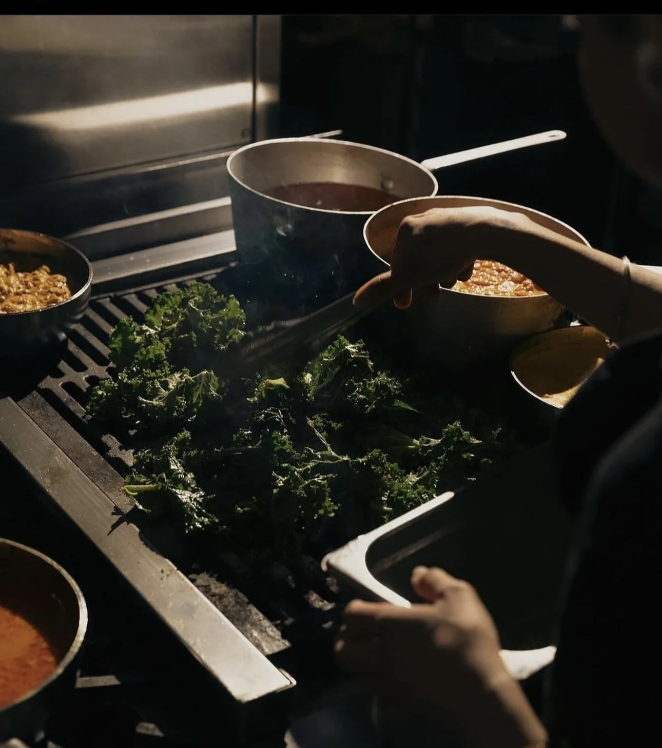Person cooking or serving food on a stove with leafy greens and various dishes in bowls.