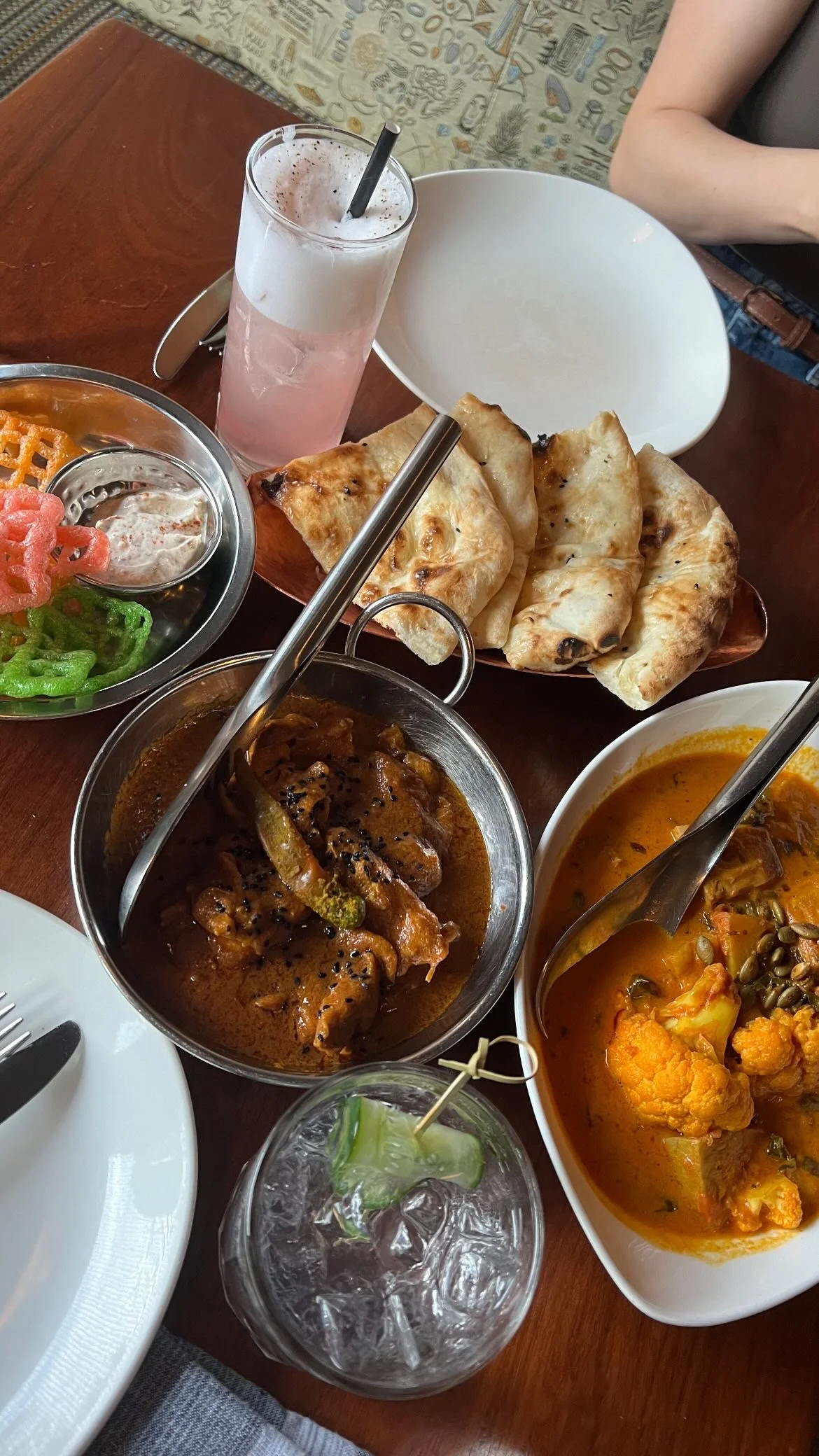A variety of Indian dishes on a wooden table, including naan bread, curry dishes, a glass of pink drink with a straw, a glass of clear beverage with ice and cucumber, and a small plate with colorful green and red snacks.