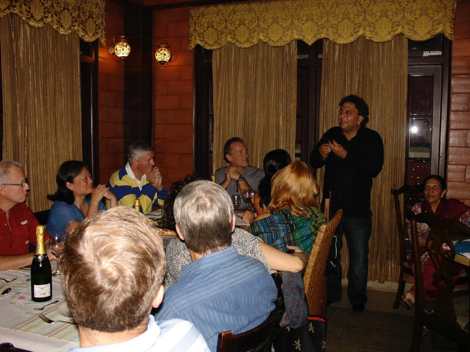 A man is giving a speech to a group of people seated around a dining table in a restaurant. The audience appears engaged, listening attentively or smiling. The setting features warm wooden walls, patterned curtains, and hanging lantern lights.