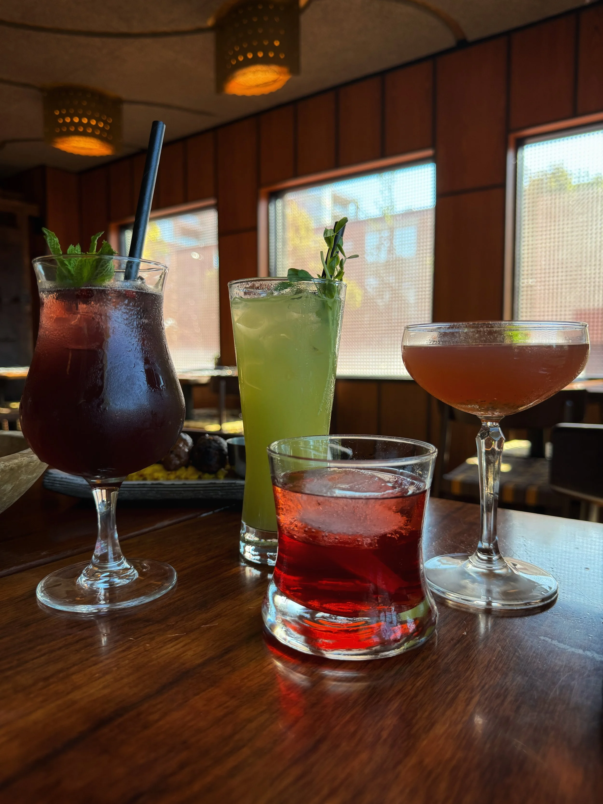 Four colorful cocktails on a wooden table inside a restaurant, with large windows in the background.